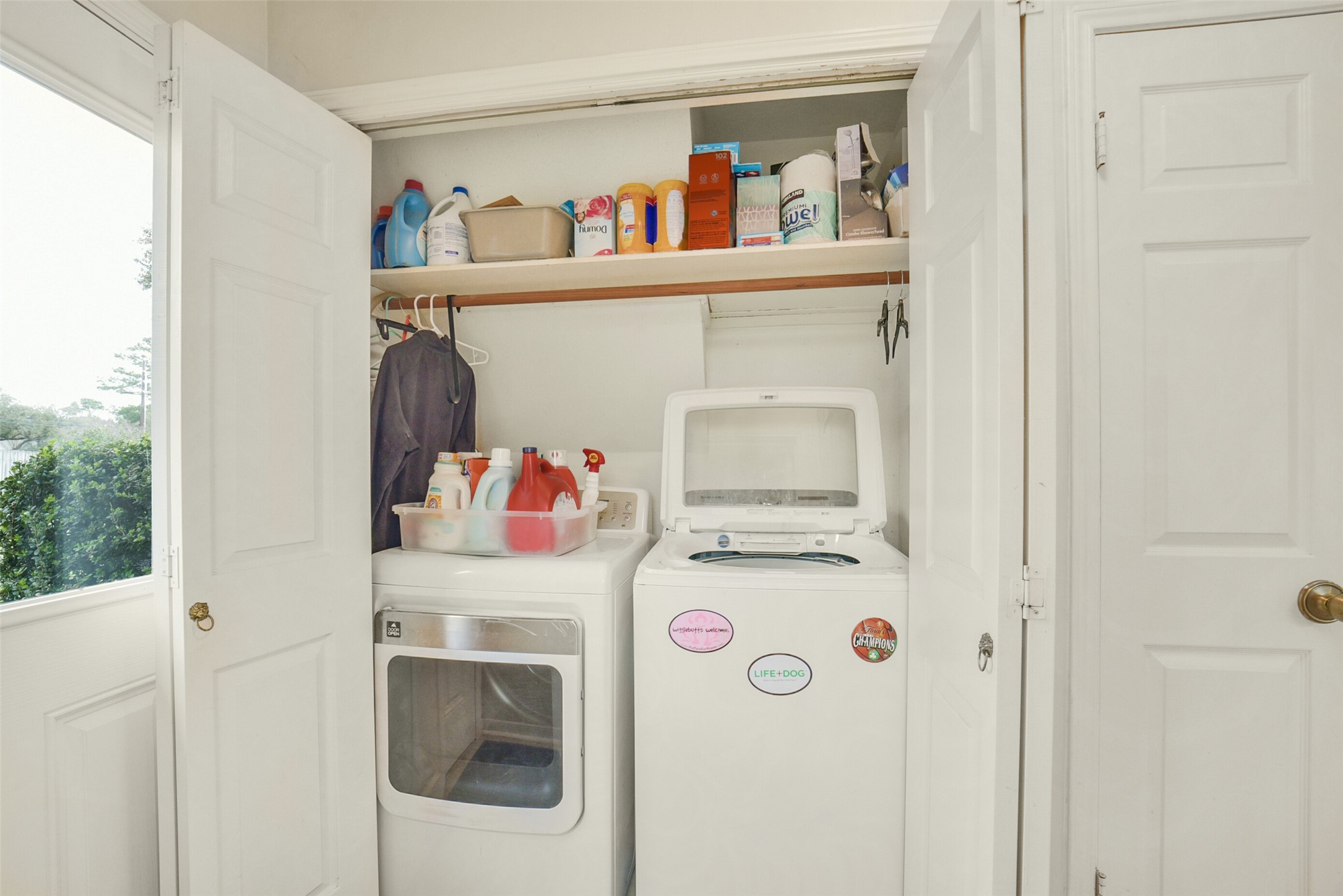 10410 Dude Road Houston, TX 77064 - Photo 23 of 45 This photo shows a compact laundry area with a washer and dryer tucked into a closet space. It features shelving above for storage of laundry essentials and cleaning supplies. The area is well-organized and utilizes space efficiently.