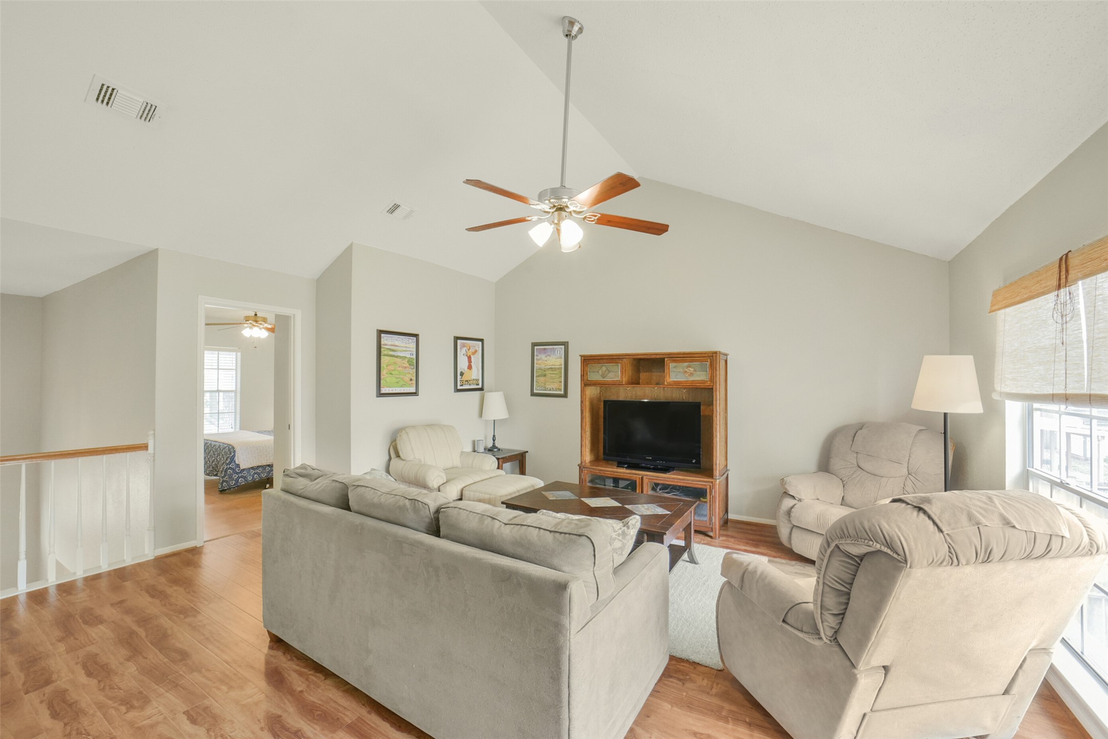 10410 Dude Road Houston, TX 77064 - Photo 25 of 45 This living room features a cozy seating area with plush sofas, a TV, and a ceiling fan. The space is well-lit with natural light from a large window, and has warm wood flooring. A bedroom is visible through an adjacent doorway.