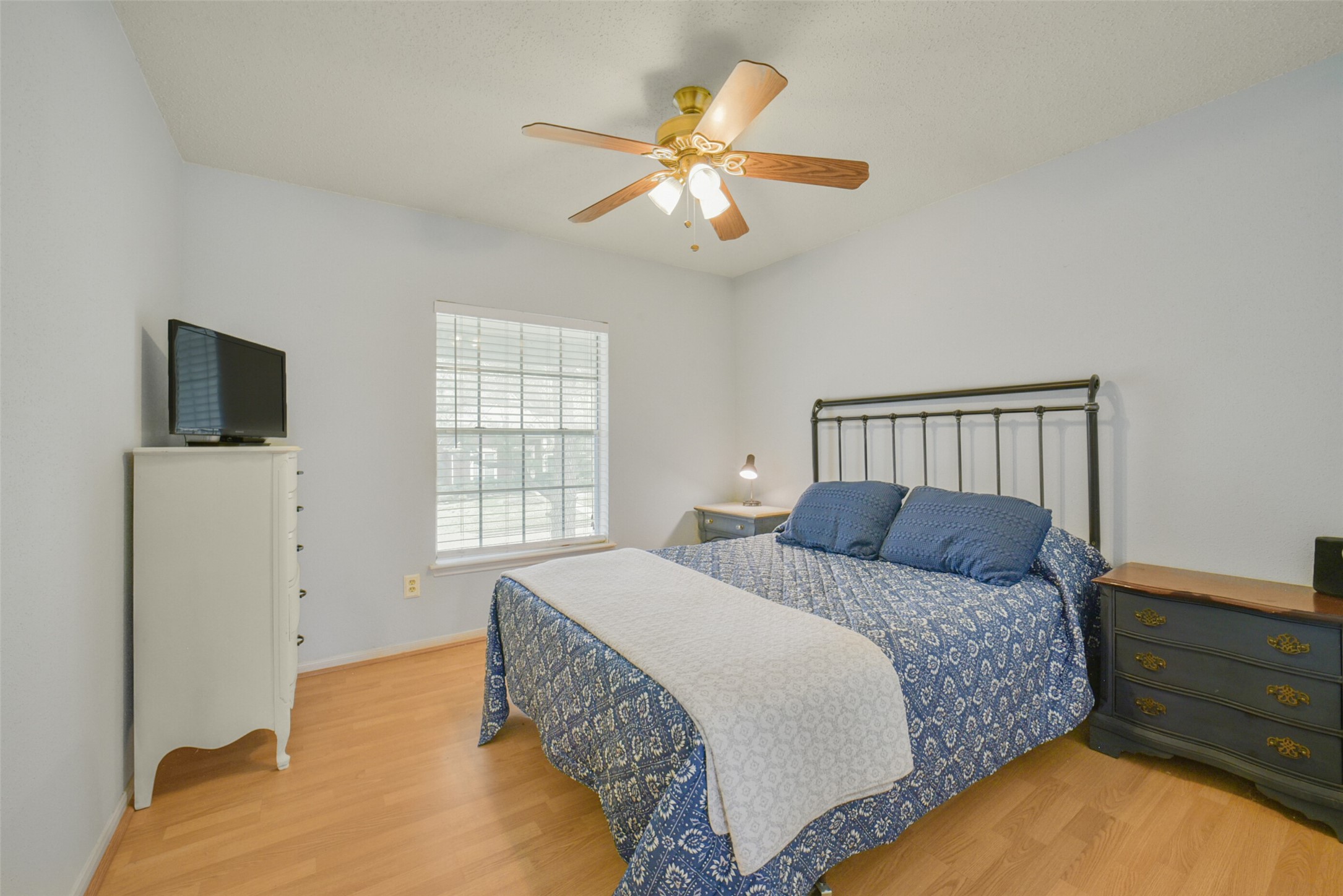 10410 Dude Road Houston, TX 77064 - Photo 27 of 45 This cozy bedroom features light blue walls and wooden flooring, with a large window that lets in natural light. It includes a ceiling fan, a bed with a dark metal frame, a chest of drawers with a TV on top, and a side table with a lamp, creating a comfortable and functional space.