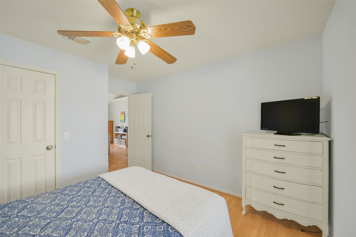 10410 Dude Road Houston, TX 77064 - Photo 28 of 45 This cozy bedroom features soft blue walls, a ceiling fan, and hardwood floors. It includes a white dresser with a TV and an inviting bed with a patterned blue bedspread. There's a glimpse into an adjacent room through the open door.