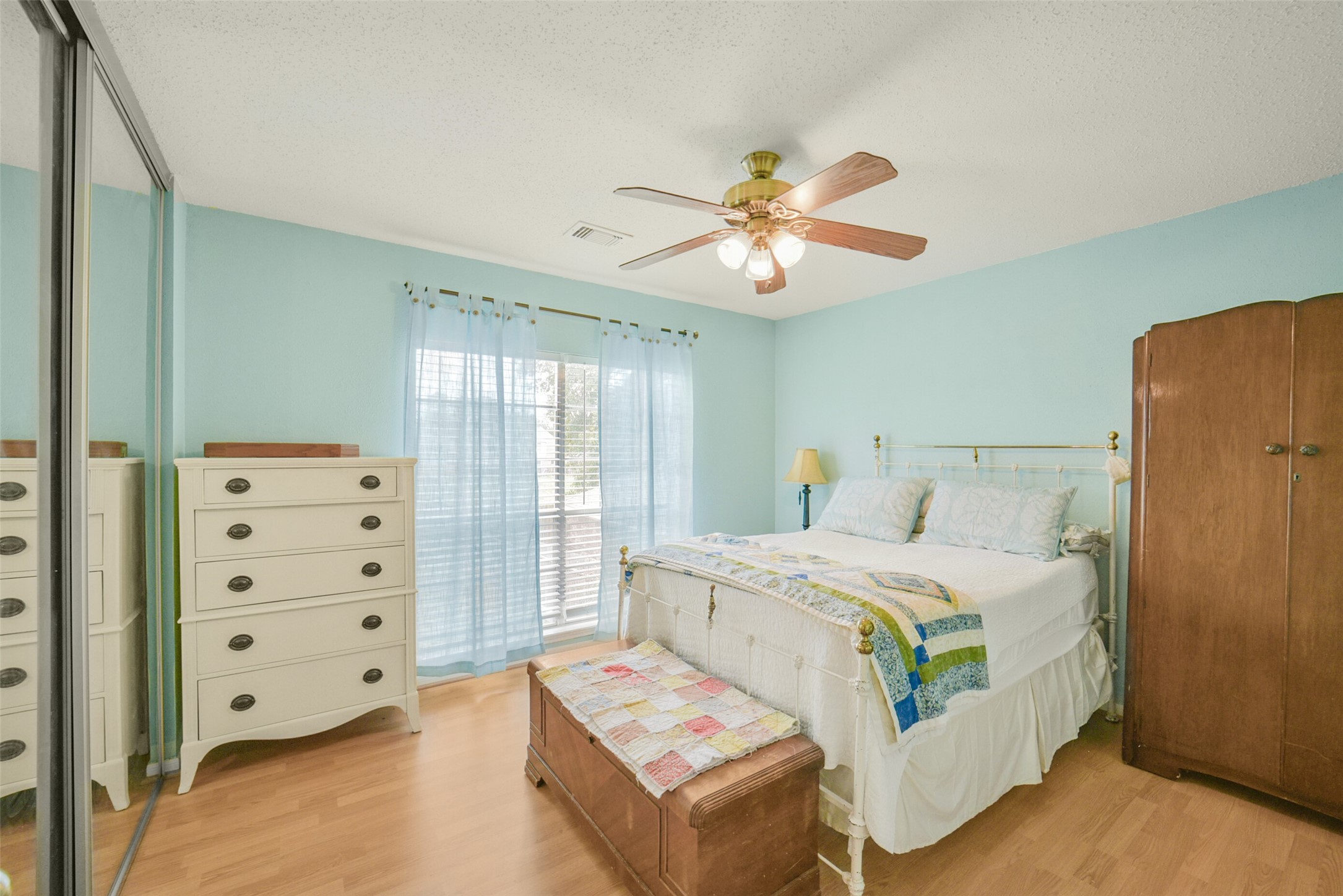 10410 Dude Road Houston, TX 77064 - Photo 29 of 45 This bedroom features soft blue walls, a ceiling fan, large mirrored closet doors, and ample natural light from a window with sheer curtains. It includes a cozy bed, a wooden wardrobe, a chest of drawers, and a colorful quilt, creating a warm and inviting atmosphere.
