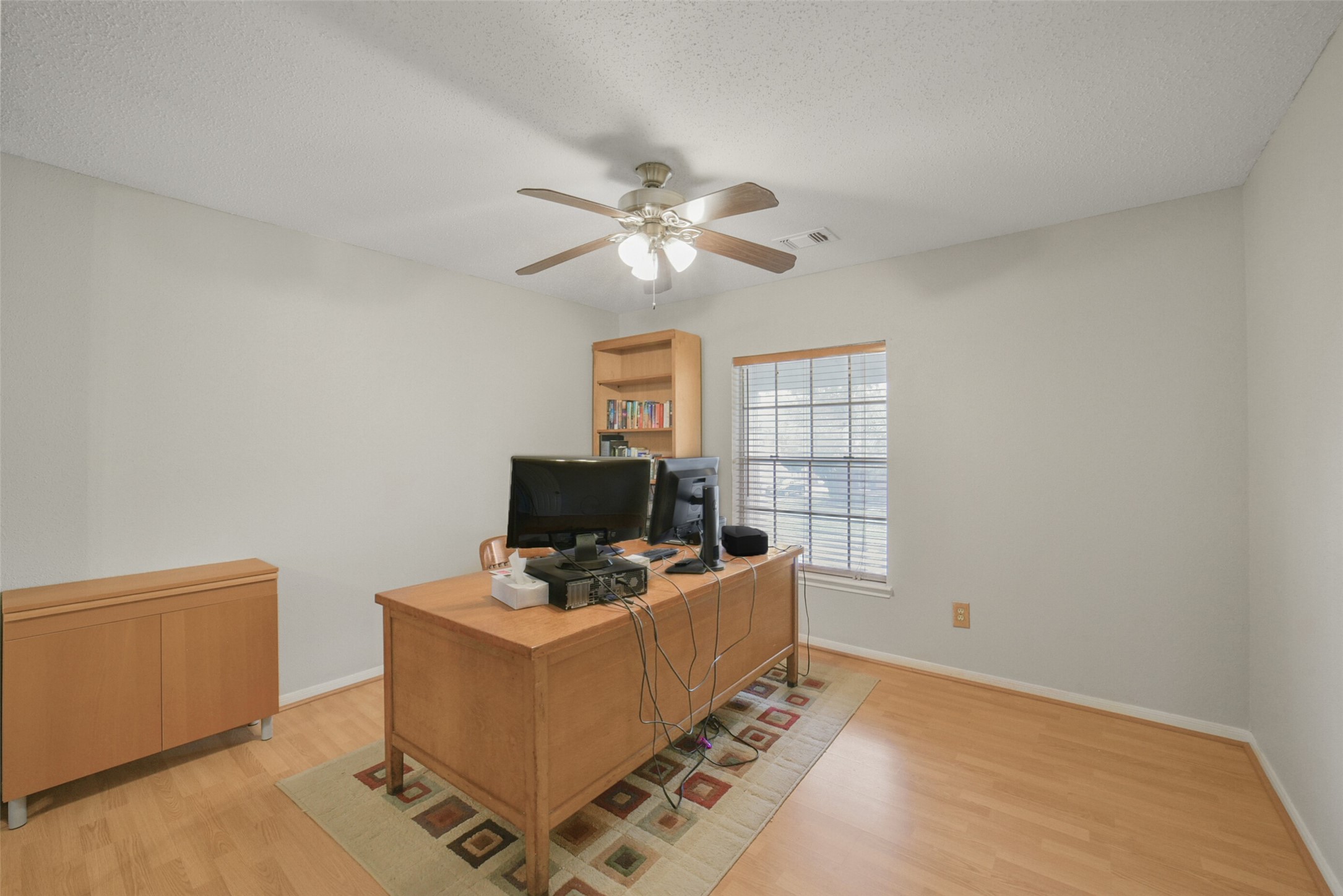 10410 Dude Road Houston, TX 77064 - Photo 32 of 45 This photo shows a bright home office with a ceiling fan, light wood flooring, and a large window for natural light. It includes a desk with a computer setup and a bookshelf, creating a functional workspace.