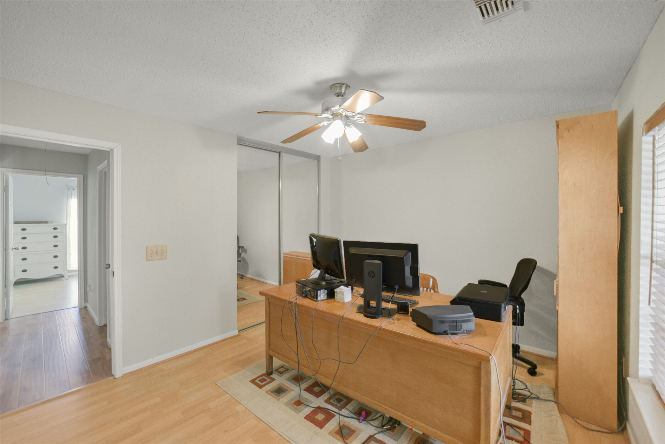 10410 Dude Road Houston, TX 77064 - Photo 33 of 45 This photo shows a well-lit home office with a ceiling fan, wooden desk, computers, and a printer. The room has light-colored walls, a mirrored closet, and hardwood floors, offering a cozy workspace.