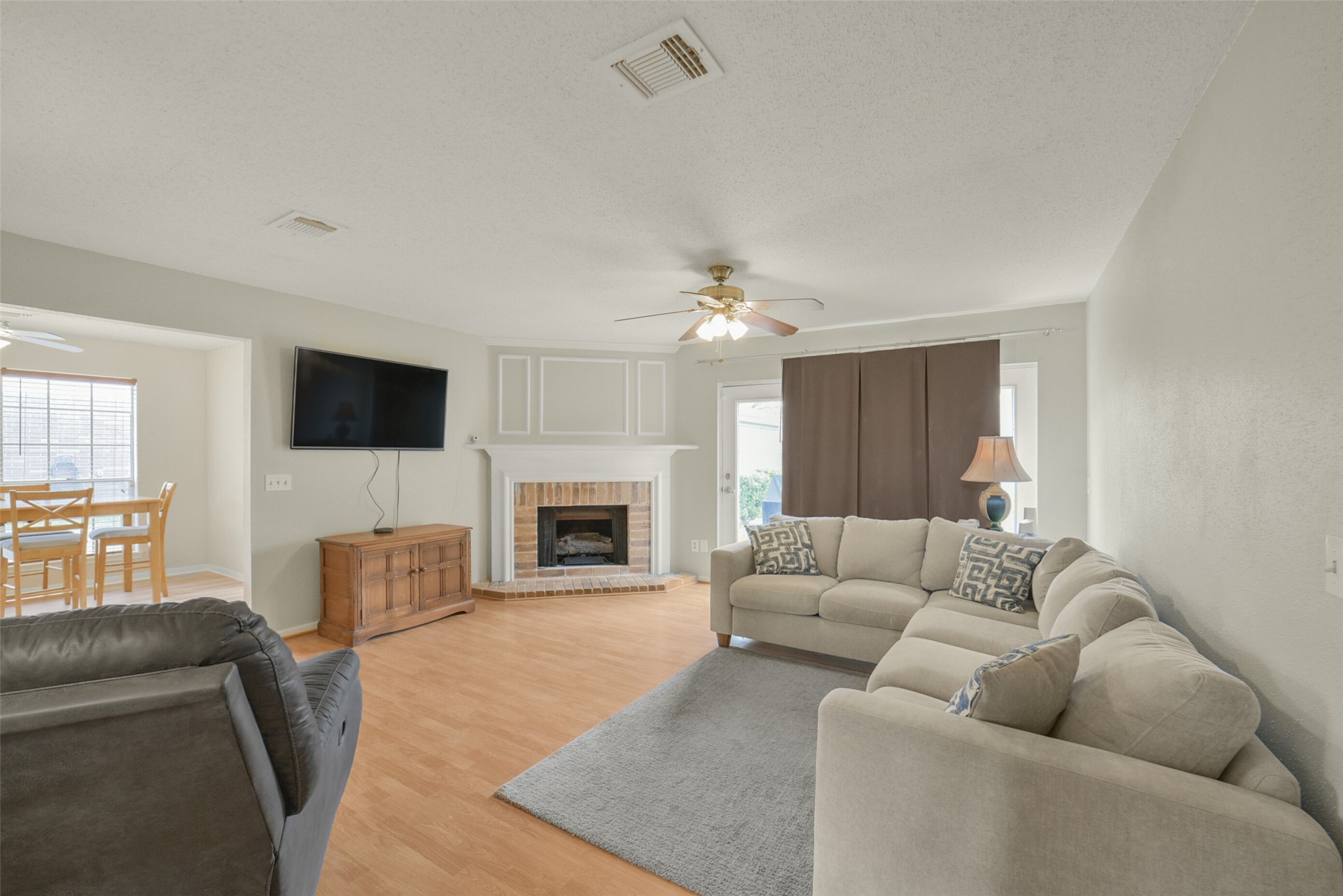 10410 Dude Road Houston, TX 77064 - Photo 4 of 45 Bright and spacious living room with a cozy fireplace, ceiling fan, and mounted TV. The room features a comfortable sectional sofa, a neutral color palette, and laminate flooring. Adjacent to the dining area, offering a seamless flow for entertaining.