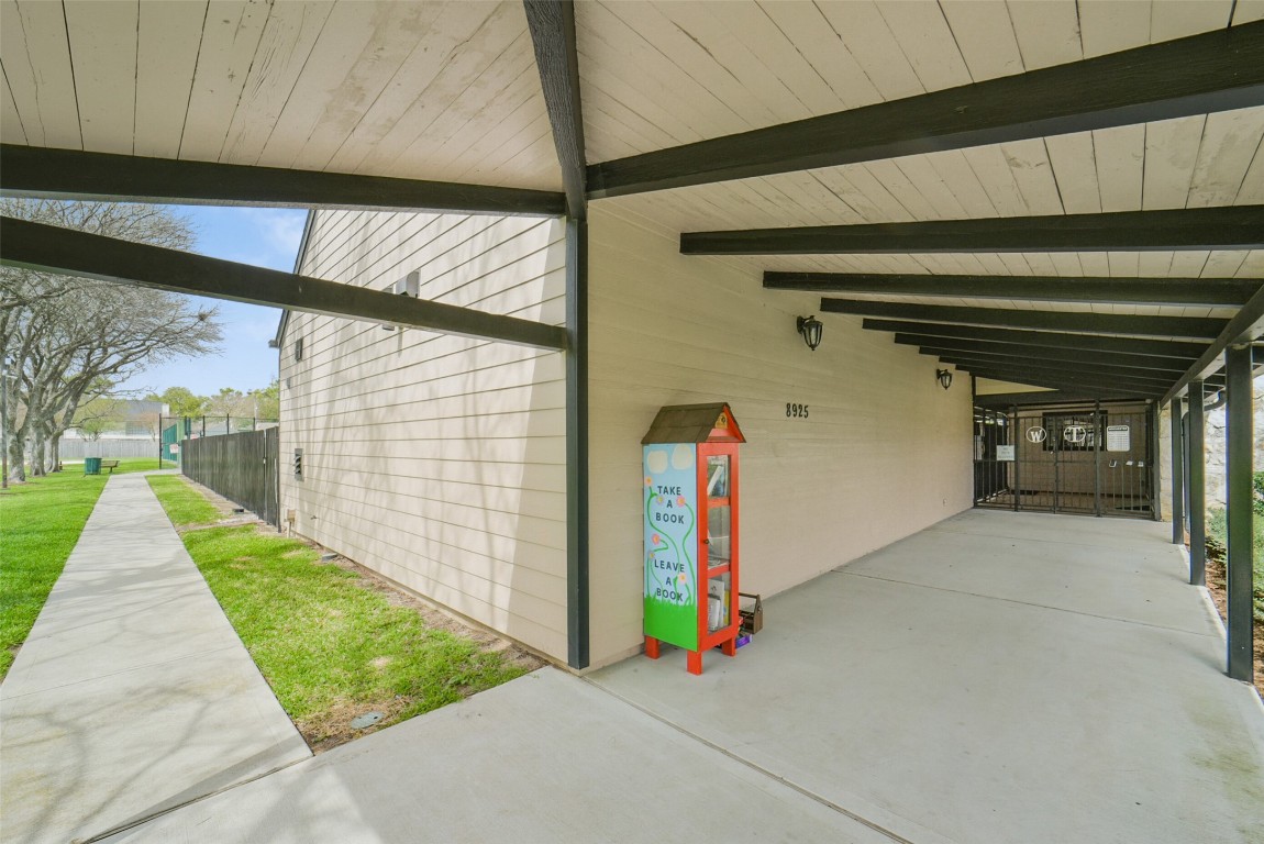 10410 Dude Road Houston, TX 77064 - Photo 41 of 45 This photo shows a covered walkway with a modern, wooden exterior. There's a small, colorful book exchange box labeled "Take a book, leave a book," adding a community-friendly touch. The area is well-maintained, with a concrete path leading to a fenced outdoor space and greenery alongside.
