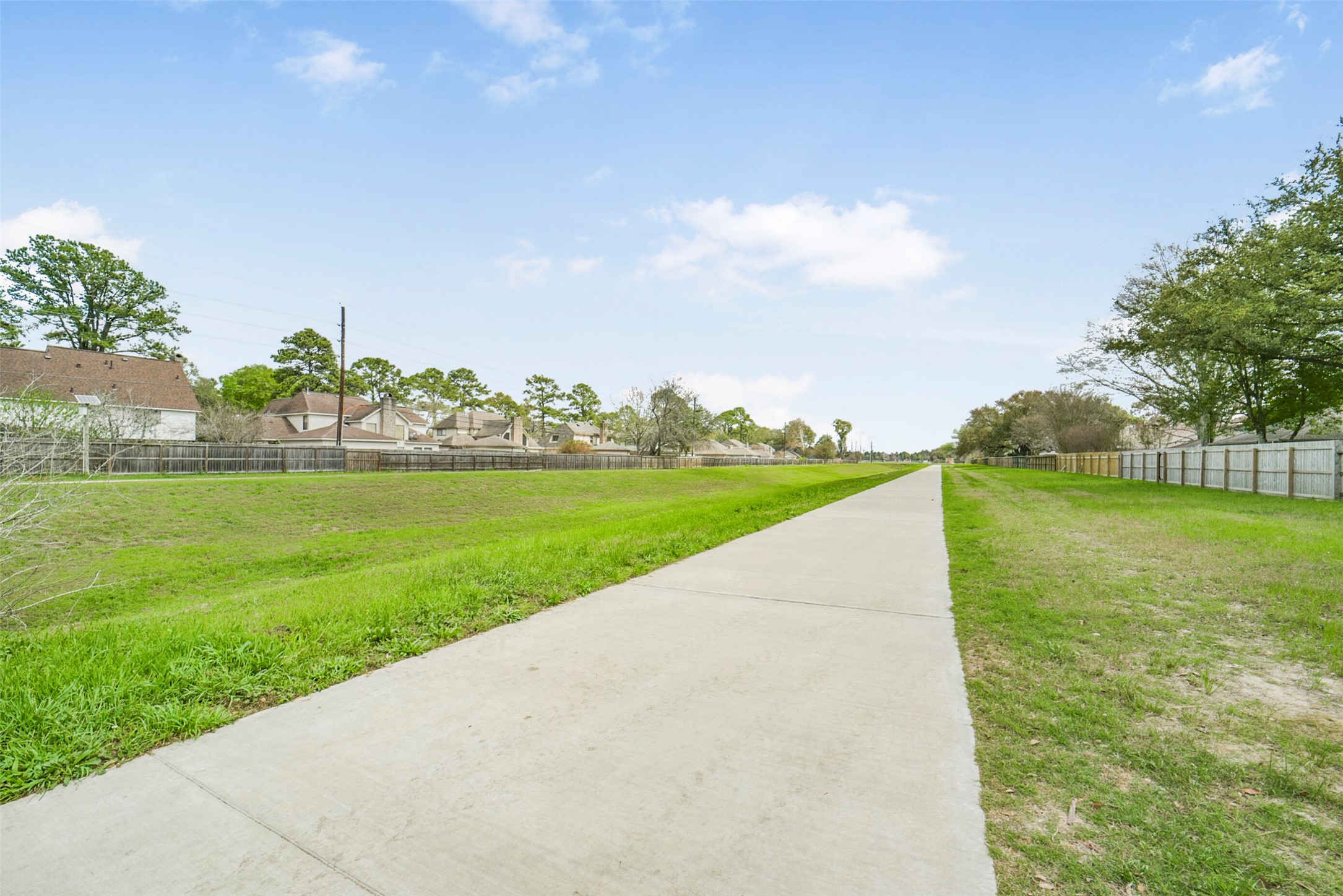 10410 Dude Road Houston, TX 77064 - Photo 44 of 45 This photo showcases a long, paved walking path with grassy areas on both sides, leading through a residential neighborhood. The area is lined with fences and mature trees, offering a serene and inviting environment.