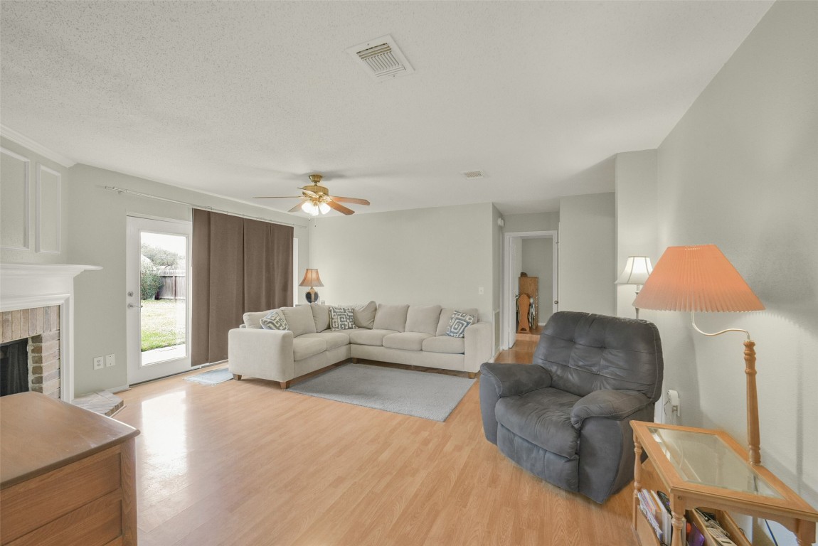 10410 Dude Road Houston, TX 77064 - Photo 5 of 45 Bright and inviting living room featuring laminate wood flooring, a cozy fireplace, and a large sectional sofa. The space is enhanced by natural light from a glass door leading to the backyard, and complemented by a ceiling fan and warm lighting.