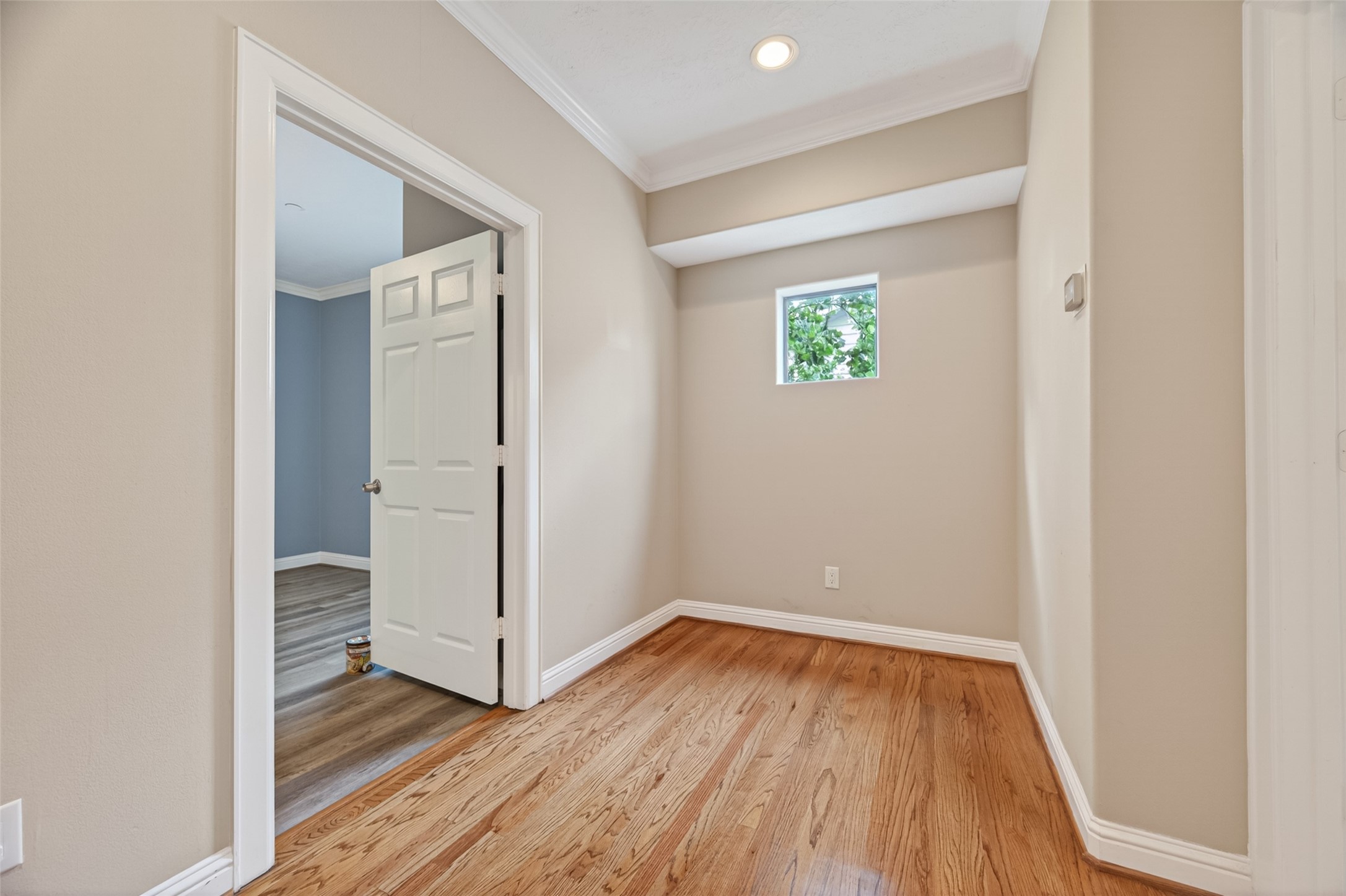 2611 Riverside Drive, Unit C Houston, TX 77004 - Photo 28 of 50 This photo showcases a small hallway with light wood flooring and beige walls. It features a recessed ceiling light and a small window providing natural light. An open door leads to an adjacent room with darker wood flooring and blue-gray walls.