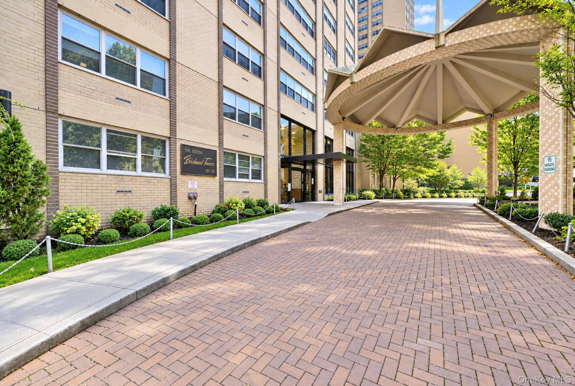 102-30 66th Road, Unit 17E Queens, NY 11375 - Photo 1 of 21 Brick-patterned driveway leading to a covered entrance with a multi-paneled canopy, featuring a brick facade building and structured landscaping