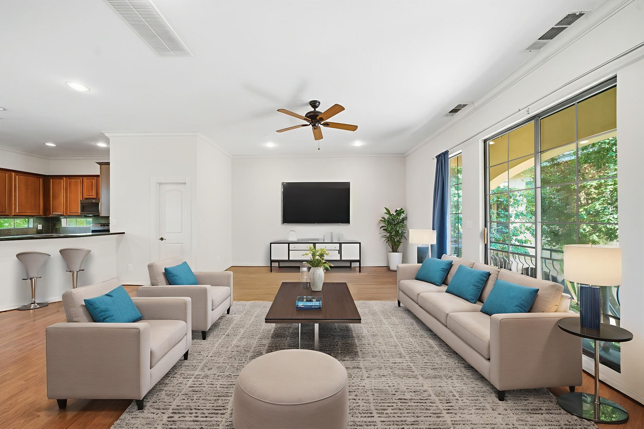 This is a bright, spacious living area featuring modern furniture and decor. It has an open layout with a view into the kitchen, light wood flooring, a ceiling fan, and large windows that allow natural light to fill the space. Digitally Staged