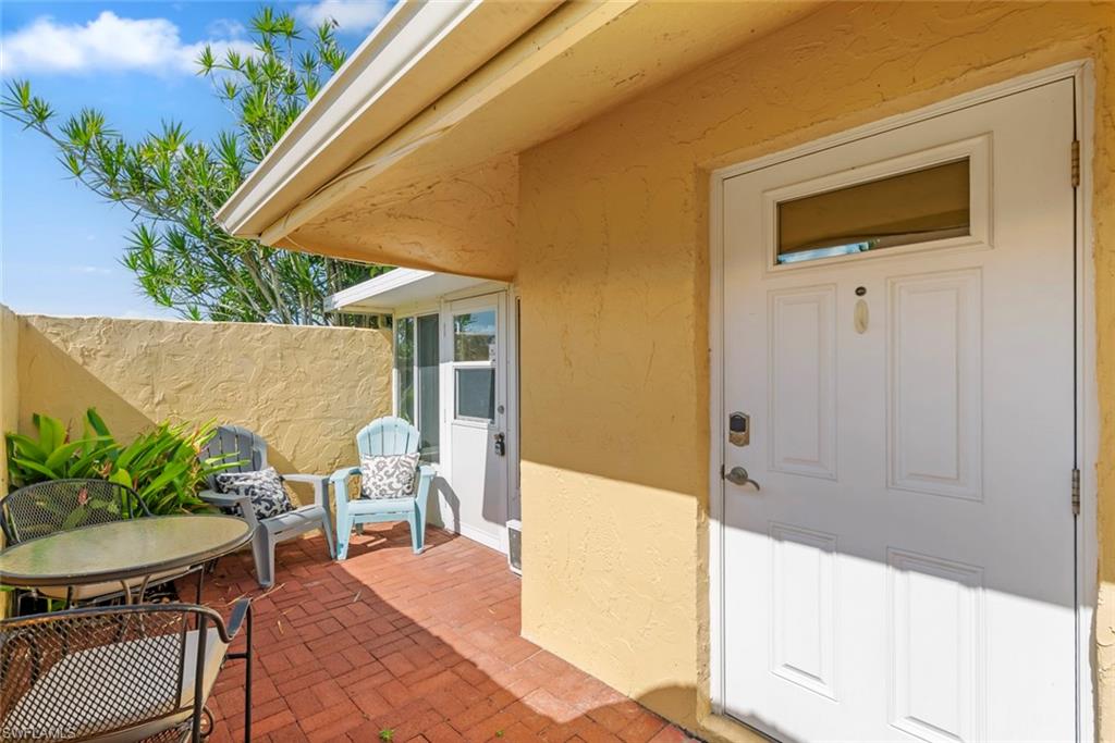136 Harrison Road, Unit 1 Naples, FL 34112 - Photo 16 of 29 Inviting entry patio featuring a white paneled door with transom window, textured stucco exterior, red brick paving, and a garden bed with lush foliage