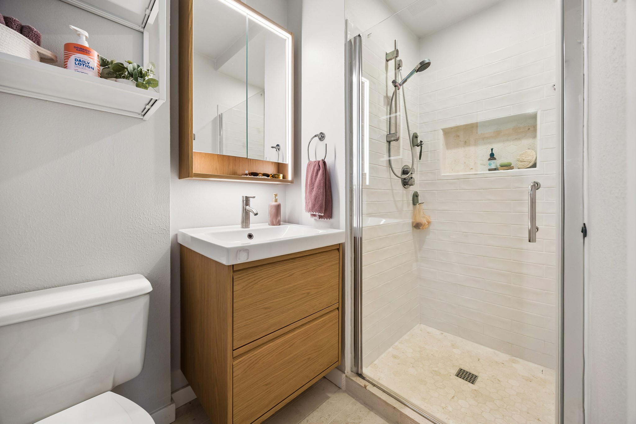 4514 Warm Springs Road Houston, TX 77035 - Photo 22 of 31 The completely renovated primary bathroom features a sleek glass-enclosed shower with a built-in niche, a floating wood vanity with an illuminated mirror that doubles as a large medicine cabinet. Convenient open shelving above the toilet and towel hooks on the wall to the right offers efficient use of space. The neutral tones and clean lines create a fresh look.