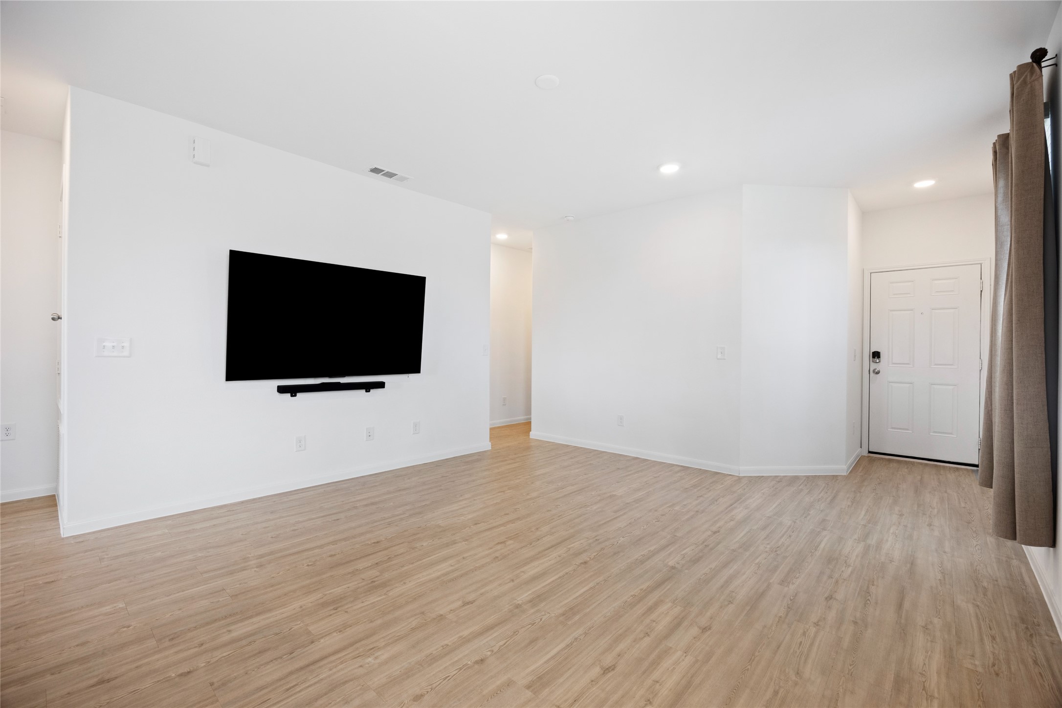 17907 Grain Auger Road Hockley, TX 77447 - Photo 10 of 24 Bright, open living space with light wood flooring, white walls, and a mounted TV. Recessed lighting adds modern touches, and a window with curtains brings in natural light. The entryway features a secure door with a keypad lock.