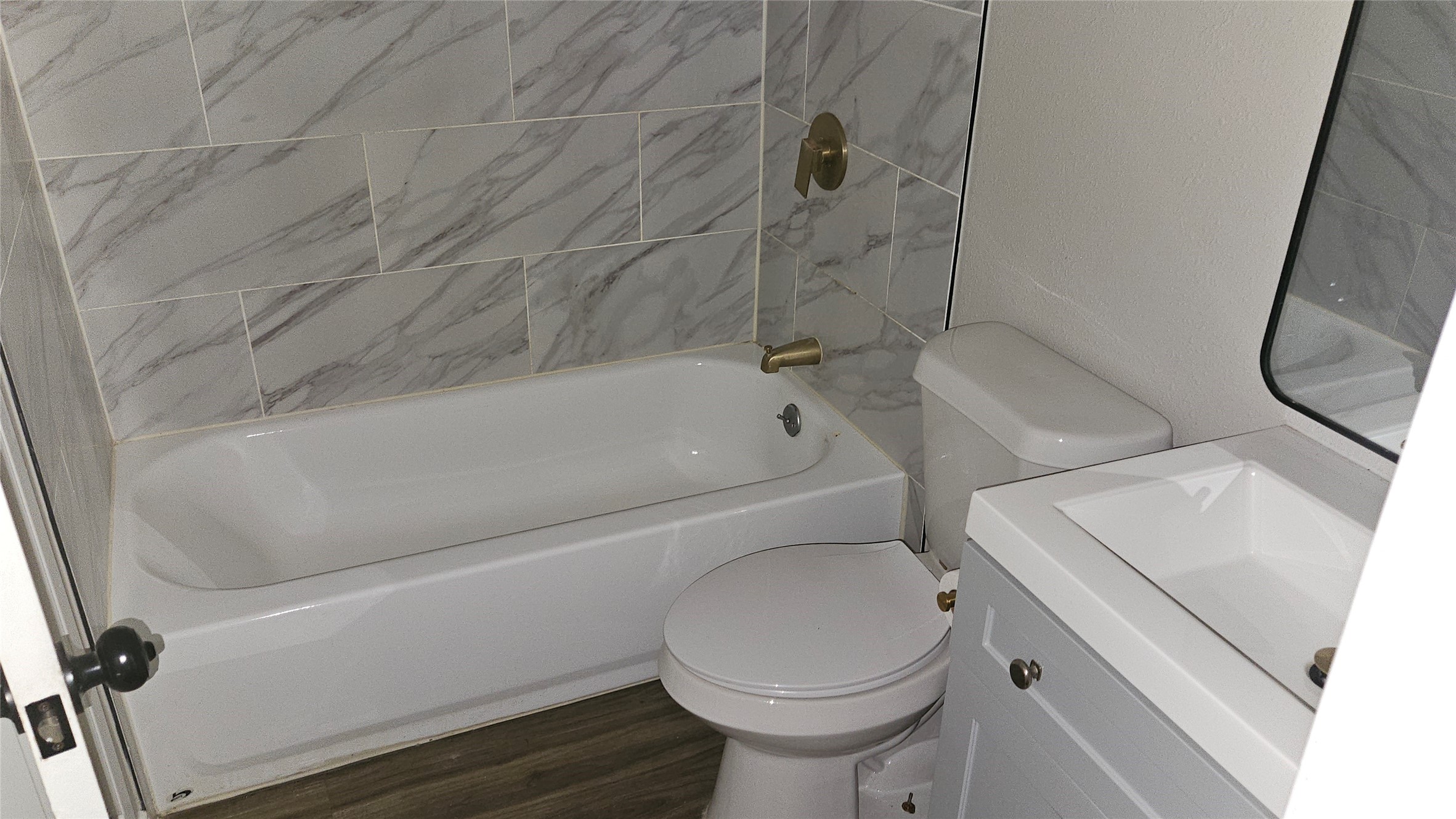 2415 Perry Street Baytown, TX 77521 - Photo 16 of 23 The primary bathroom features modern marble-style tiles, a sleek white bathtub, and matching fixtures. It includes a compact vanity with a mirror and a standard toilet, all complemented by wood-look flooring for a contemporary feel.