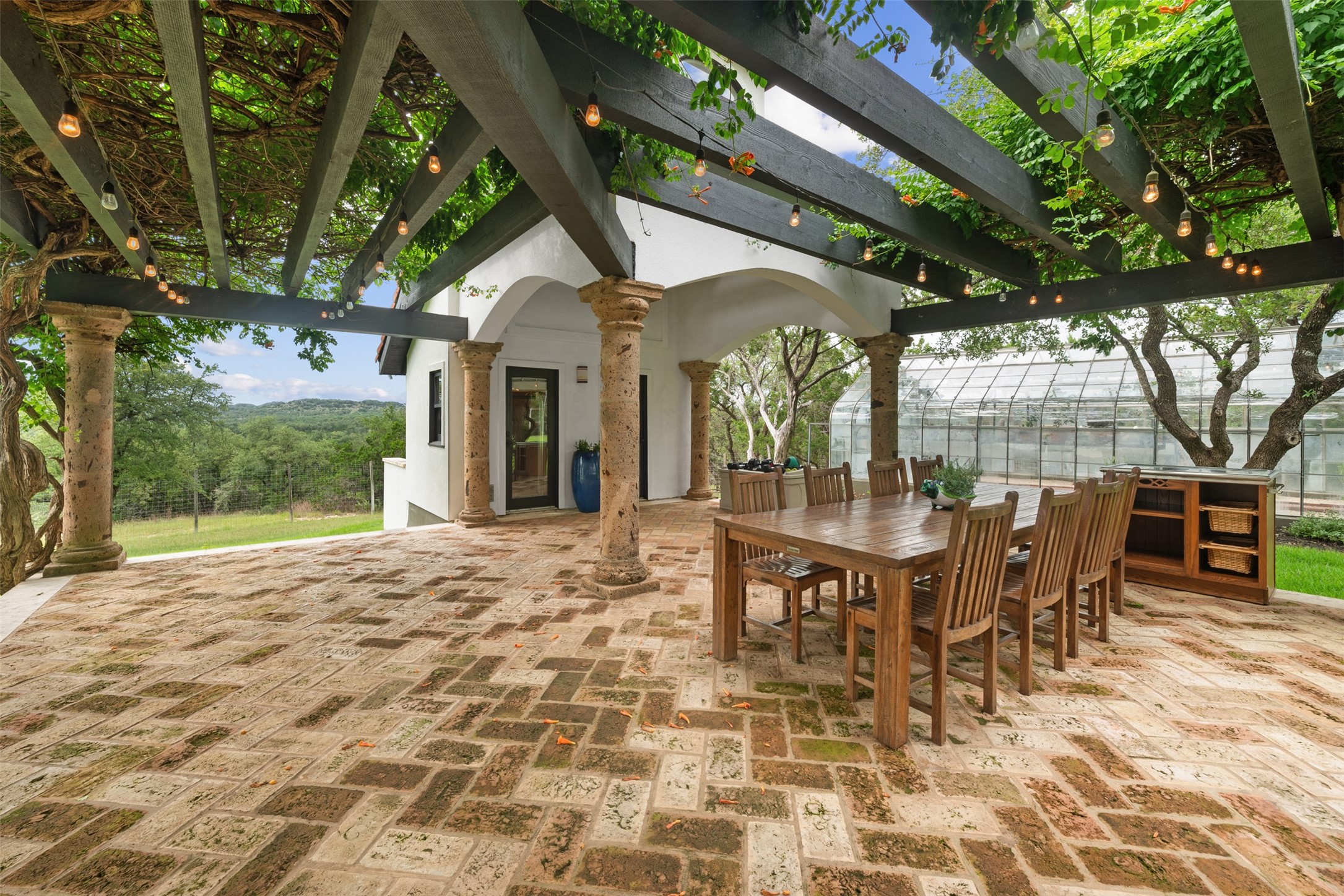 34000 Smithson Valley Road Bulverde, TX 78163 - Photo 38 of 50 Cantera stone columns, brick flooring, and overhead string lighting define this spacious covered patio designed for al fresco dining and entertaining. Surrounded by lush landscaping, it offers a private and tranquil setting.