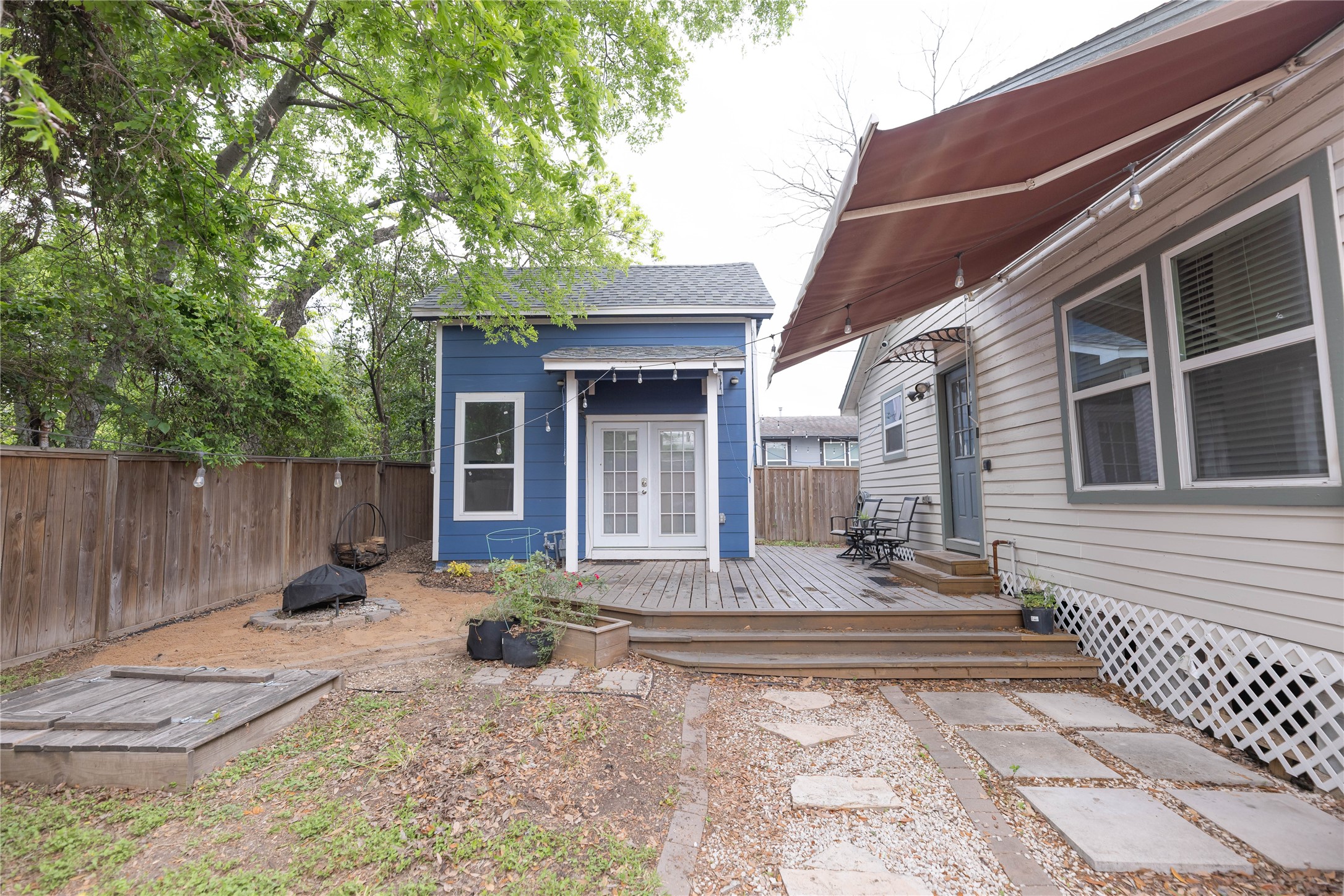 8007 Capitol Street Houston, TX 77012 - Photo 22 of 25 This cozy backyard features a charming blue outbuilding with French doors, a wooden deck, and a retractable awning. Surrounded by a wooden fence, the space includes a small patio area with stepping stones and a fire pit, offering a perfect blend of relaxation and privacy.