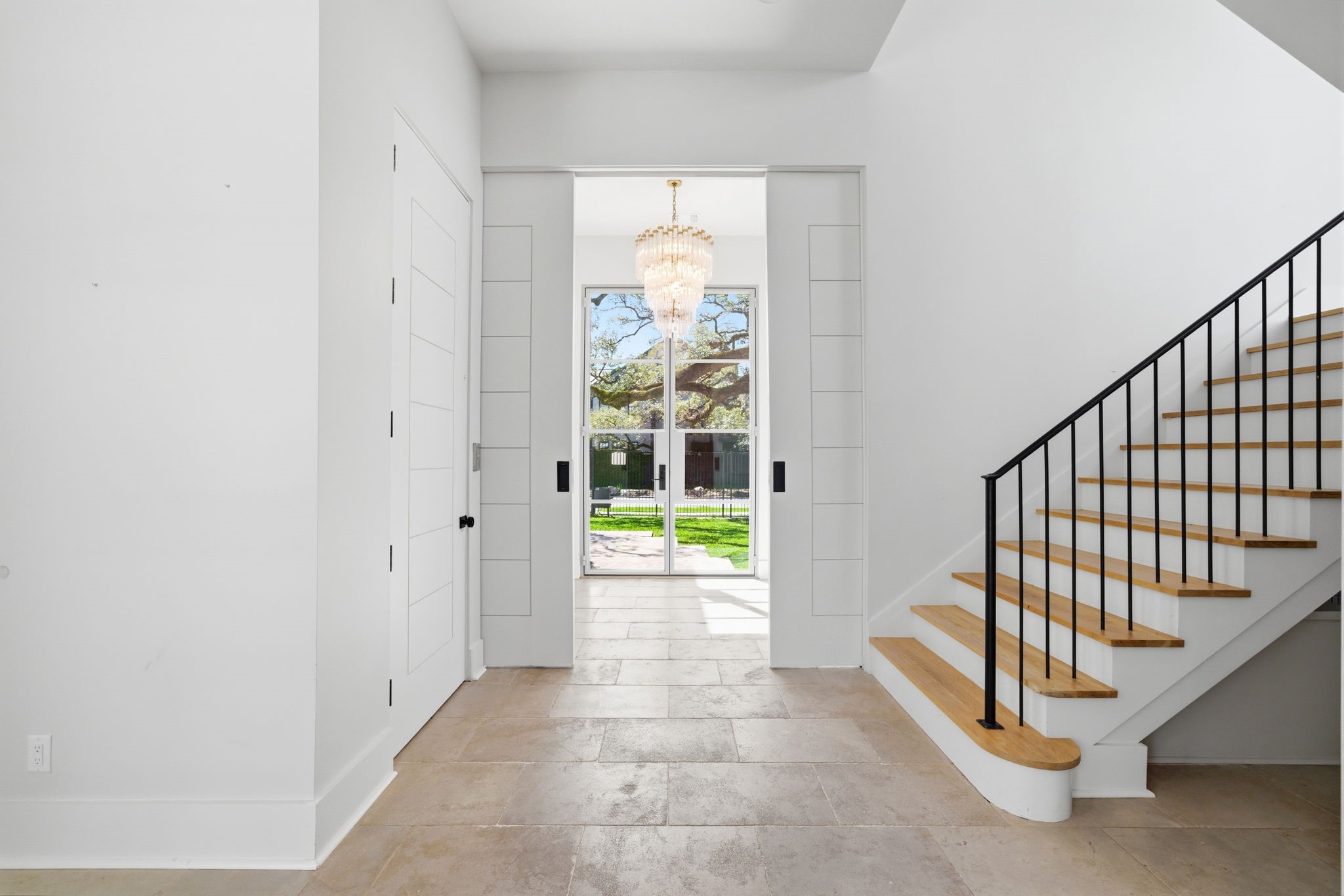2615 Yupon Street Houston, TX 77006 - Photo 11 of 50 Polished stone tile flooring and a cascading crystal chandelier define this grand foyer, which flows effortlessly into the home’s main entertainment areas. A floating wood-tread staircase with a sleek black iron railing complements the contemporary aesthetic, while pocketing doors reveal a glimpse into the dedicated study. Beyond the entry, floor-to-ceiling steel and glass doors provide immediate views of the lush courtyard and a majestic oak tree.