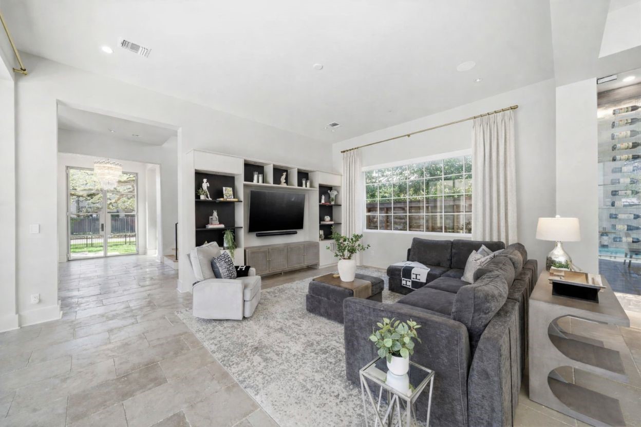 2615 Yupon Street Houston, TX 77006 - Photo 12 of 50 Spacious living room features a custom built-in media center with contrasting black accents and sleek display shelving. Large gridded windows provide views of the backyard oasis and private pool, while the open-concept layout flows toward the glass-enclosed wine room. Stone tile flooring and soaring ceilings enhance the airy, contemporary feel of this primary entertainment space.