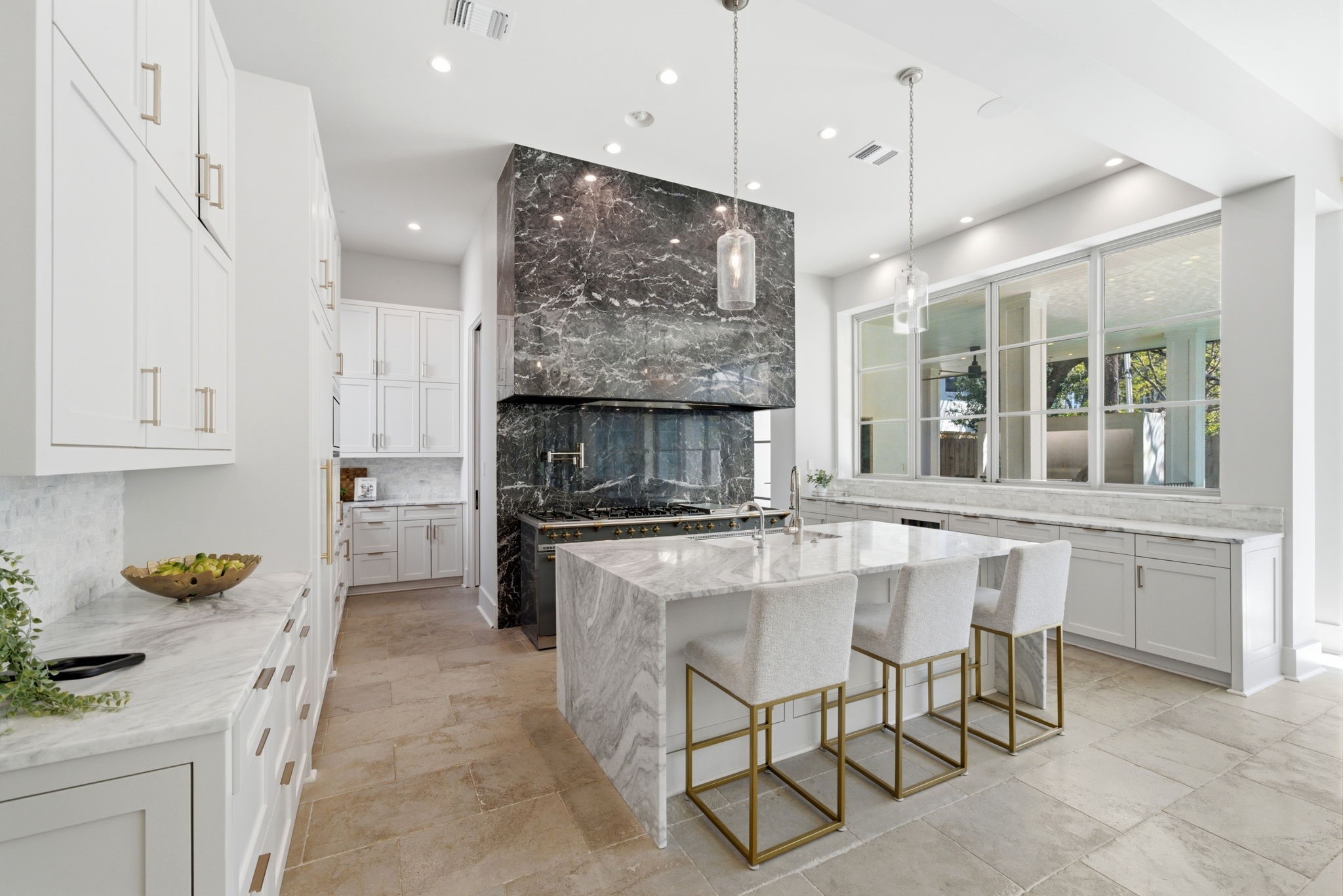 2615 Yupon Street Houston, TX 77006 - Photo 16 of 50 Professional-grade kitchen features a dramatic floor-to-ceiling black marble range hood and matching backsplash that anchors the expansive white custom cabinetry. The oversized marble waterfall island provides sophisticated counter seating, while wide gridded windows offer tranquil views of the verdant outdoor living spaces. Large-format stone tile flooring and designer pendant lighting complete this gourmet culinary retreat.