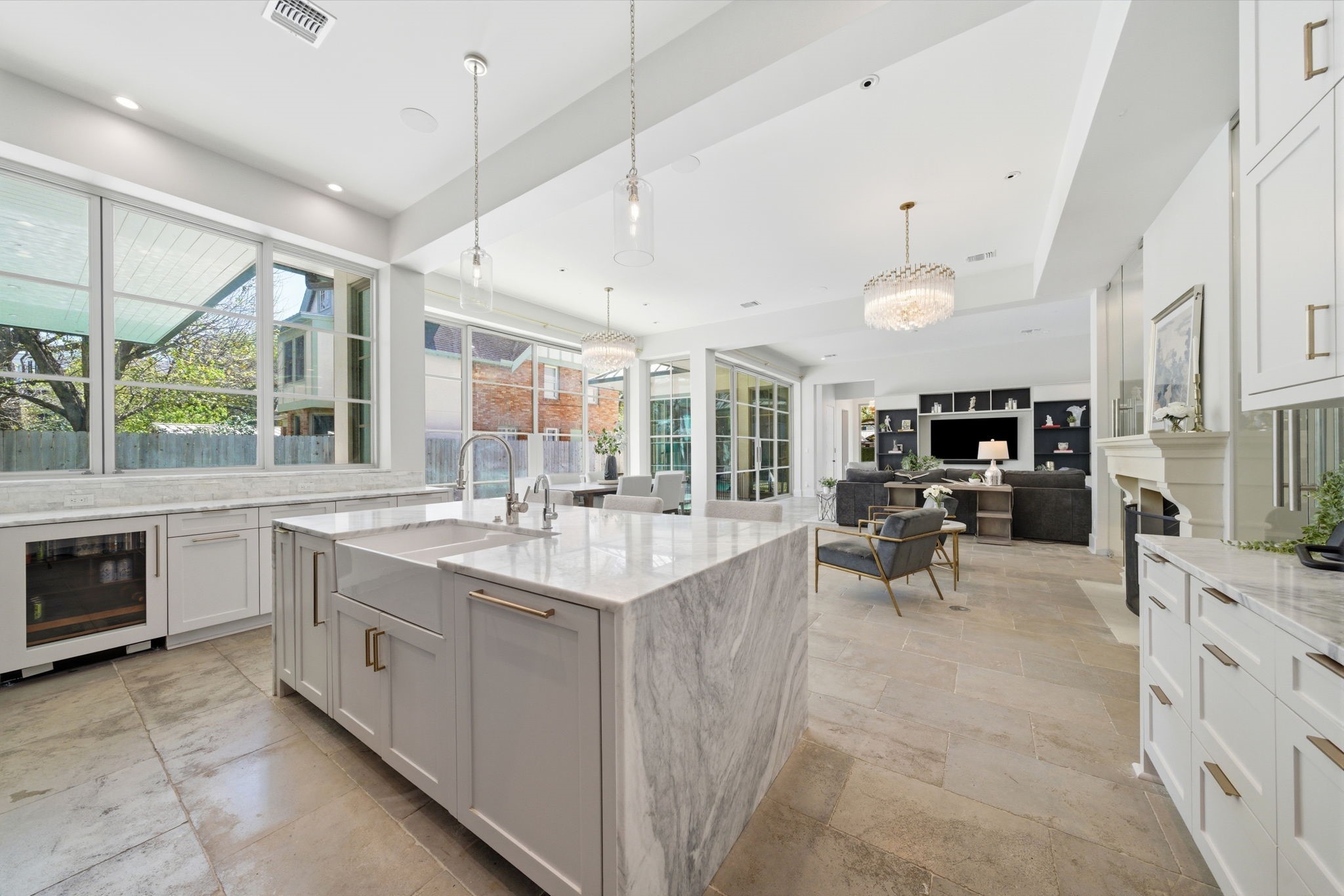 2615 Yupon Street Houston, TX 77006 - Photo 17 of 50 Stone tile flooring and soaring ceilings unify this expansive open-concept layout, seamlessly connecting the gourmet kitchen to the light-filled living area. The oversized marble waterfall island serves as a sophisticated centerpiece, featuring a farmhouse sink and professional-grade fixtures beneath designer pendant lighting. Wide gridded windows provide tranquil views of the courtyard and lush greenery, while the adjacent living room offers custom built-ins and a glass-enclosed wine room for premier enter
