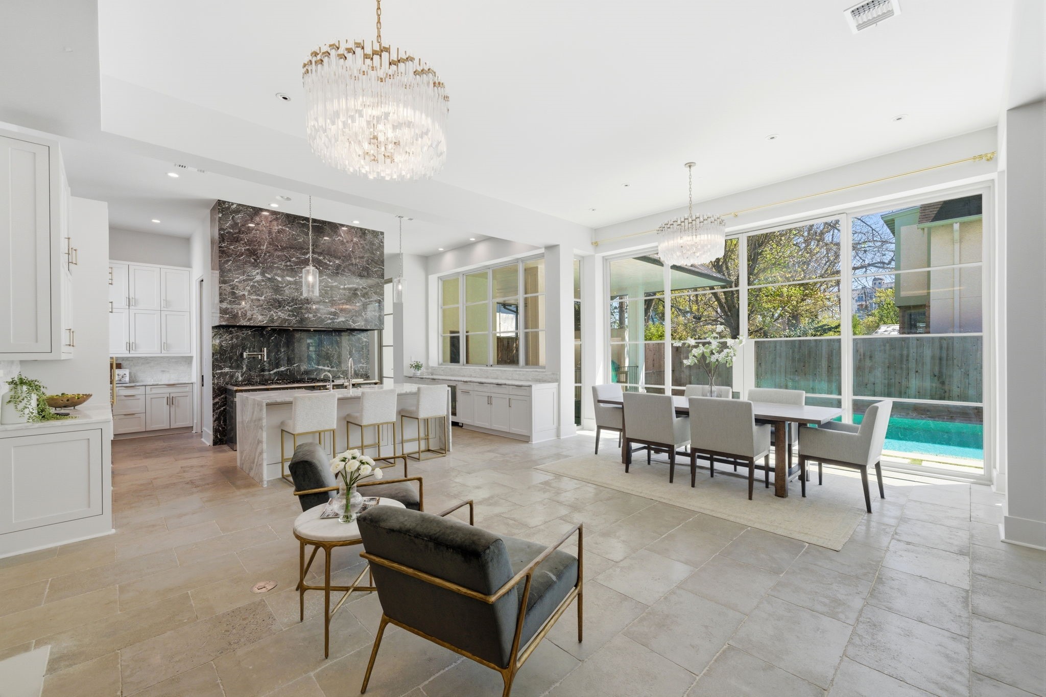 2615 Yupon Street Houston, TX 77006 - Photo 20 of 50 Large-format stone tile flooring unifies this expansive first-floor living space, where the gourmet kitchen, formal dining area, and lounge flow together effortlessly. Massive gridded windows provide a panoramic view of the private heated pool and lush backyard, flooding the room with light. Designer crystal chandeliers anchor the dining and lounge zones, while the sleek marble island and striking black stone range hood highlight the home's contemporary luxury and high-end finishes.