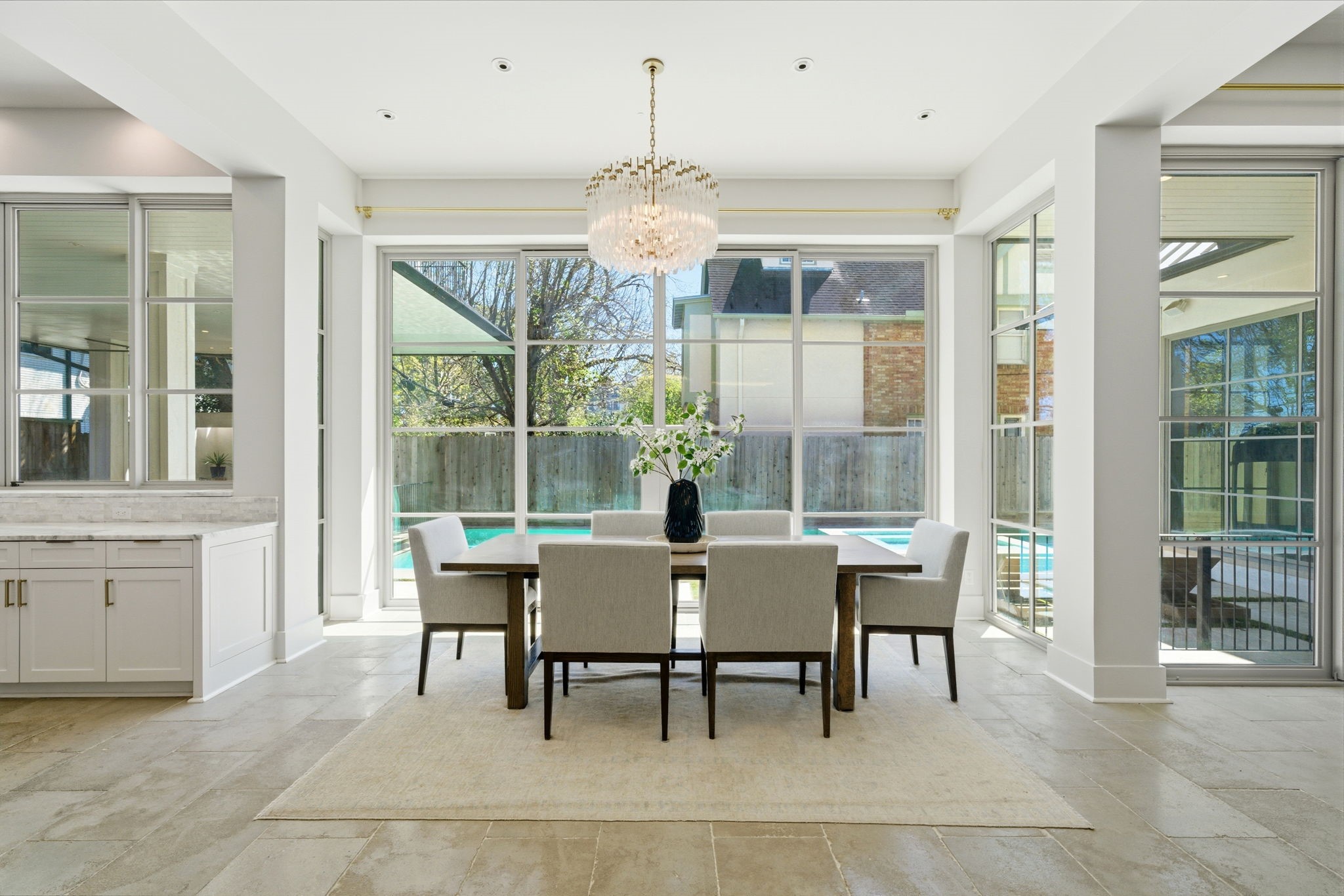 2615 Yupon Street Houston, TX 77006 - Photo 21 of 50 Floor-to-ceiling gridded windows wrap this sophisticated dining area, offering panoramic views of the heated pool and private backyard. A glamorous tiered crystal chandelier anchors the space, while a built-in buffet with marble countertops provides an elegant serving station for hosting. Stone tile flooring and an airy, open layout ensure a seamless transition to the gourmet kitchen and outdoor oasis.