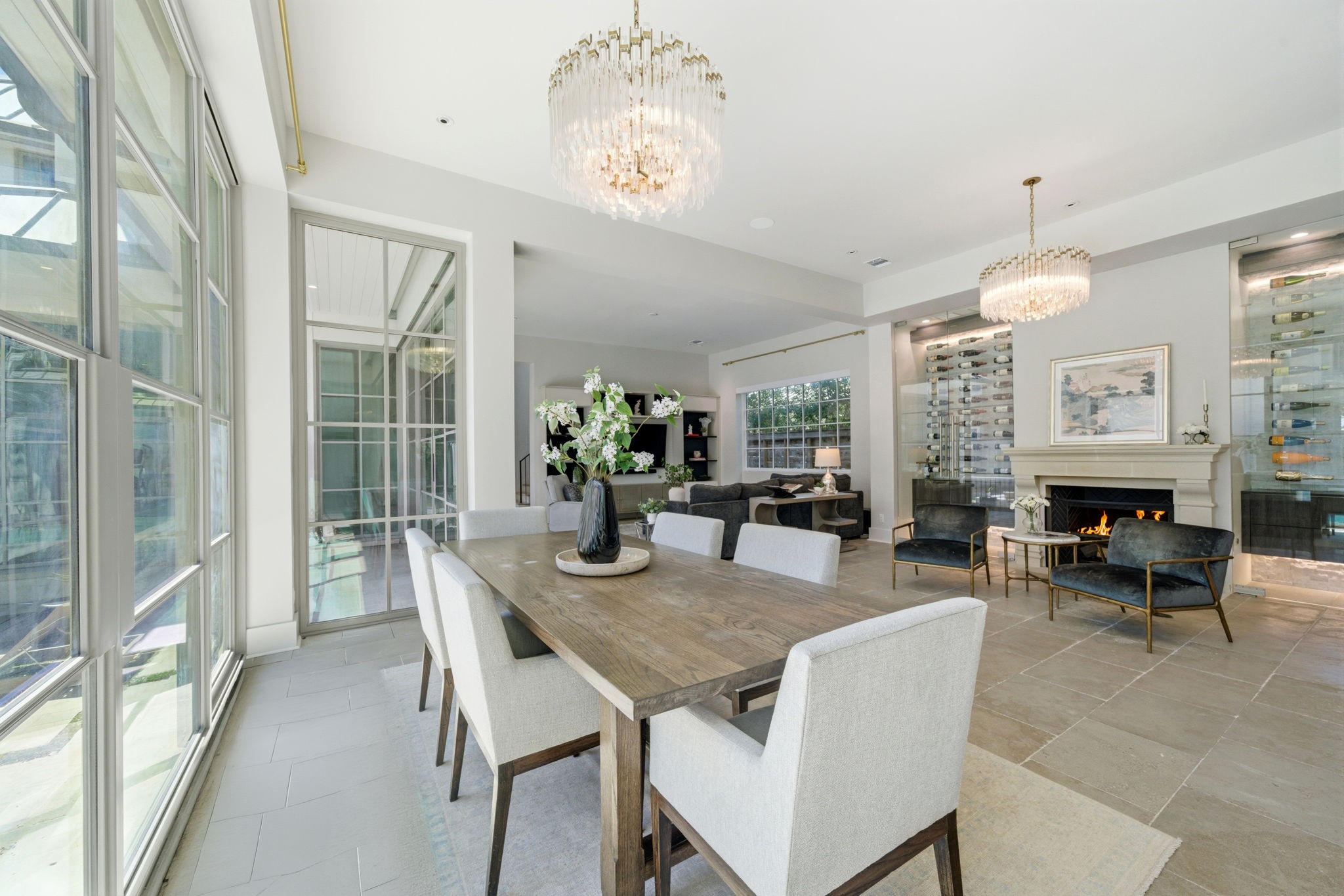 2615 Yupon Street Houston, TX 77006 - Photo 22 of 50 Sophisticated formal dining area features a large table set beneath a tiered crystal chandelier, offering views of the courtyard through floor-to-ceiling glass. A glass-enclosed wine room with wall-mounted displays and a stone-clad fireplace with contemporary velvet seating create a premier setting for upscale entertaining. Large-format stone tile floors flow throughout the open layout, seamlessly connecting this elegant space to the adjacent living room.
