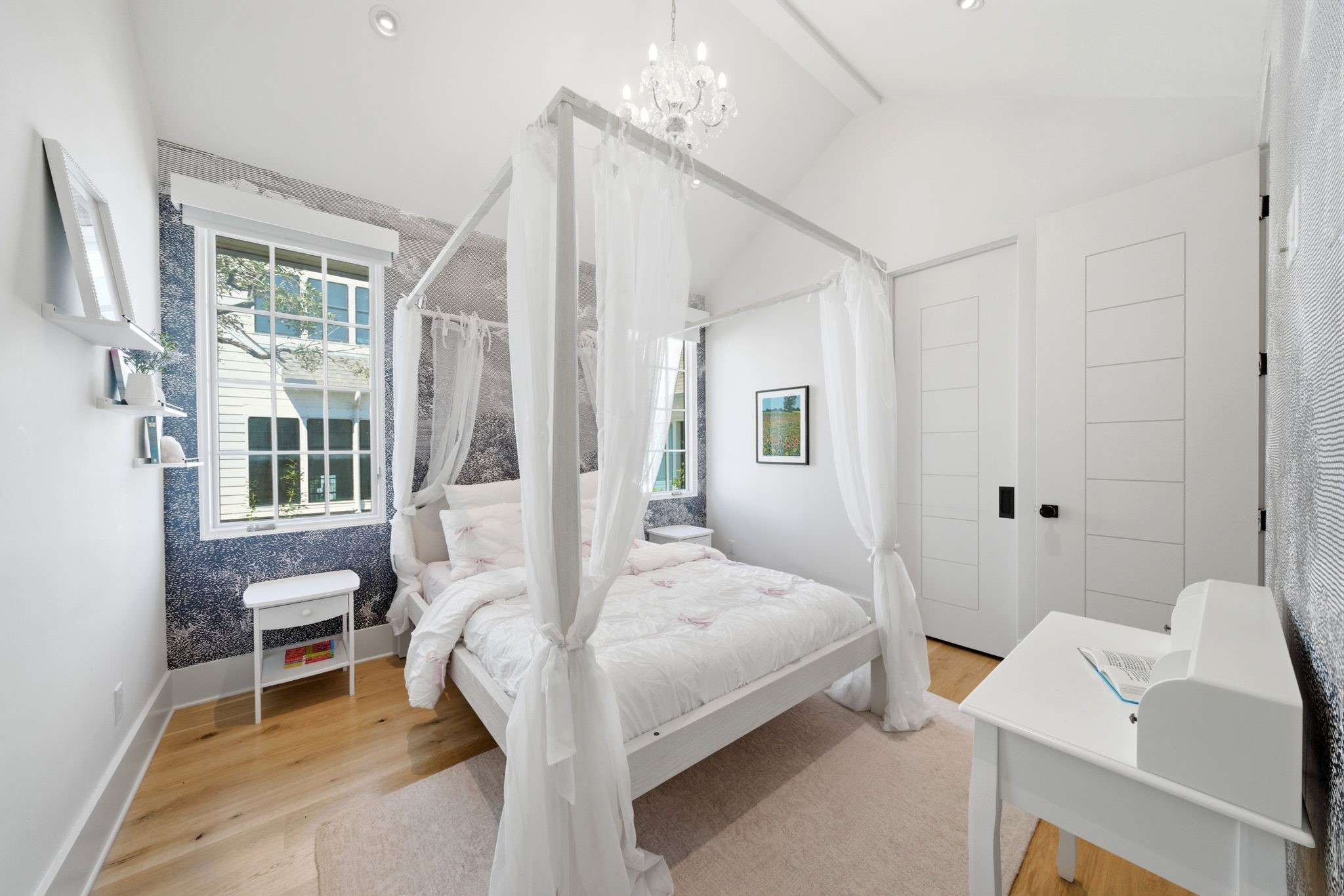 2615 Yupon Street Houston, TX 77006 - Photo 28 of 50 Vaulted ceilings and a sparkling crystal chandelier elevate this sophisticated secondary bedroom, featuring a whimsical canopy bed and a custom illustrative accent wall. Light-filled gridded windows frame views of the picturesque street, while warm white oak floors flow toward a sleek built-in desk area. This private retreat includes an ensuite bath and a large walk-in closet, blending contemporary luxury with thoughtful designer finishes.