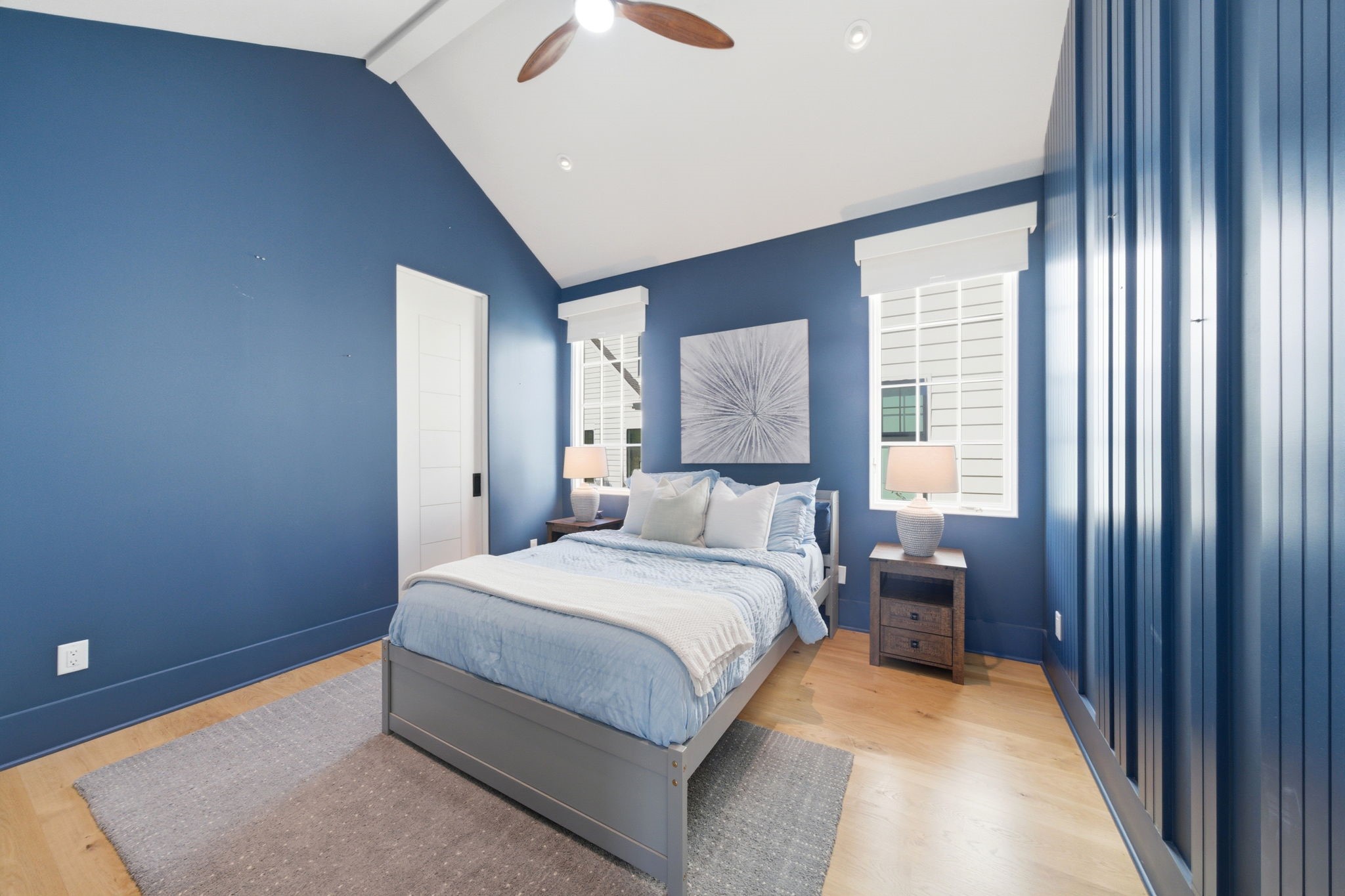 2615 Yupon Street Houston, TX 77006 - Photo 29 of 50 Vaulted ceilings and a contemporary wood-blade fan crown this vibrant secondary bedroom, anchored by a deep navy accent wall and textured vertical paneling. Large gridded windows provide ample natural light and views of the picturesque street, while wide-plank white oak floors add warmth to the designer space. This private retreat includes an ensuite bath and a large walk-in closet for ultimate comfort.