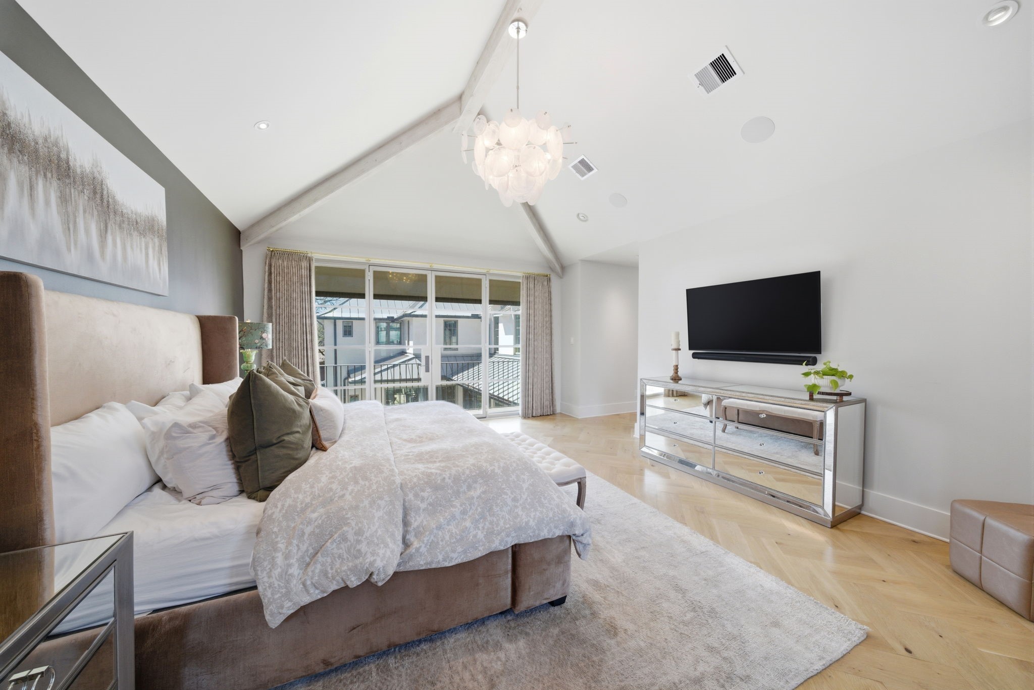 2615 Yupon Street Houston, TX 77006 - Photo 30 of 50 Vaulted ceilings and a contemporary petal-style chandelier crown this expansive primary suite, where wide-plank herringbone white oak floors add timeless luxury. Floor-to-ceiling gridded glass doors offer easy access to a private balcony overlooking the lush yard, flooding the room with light. This tranquil retreat features a minimalist wall-mounted media center and ample space for a king-sized bed and seating area.