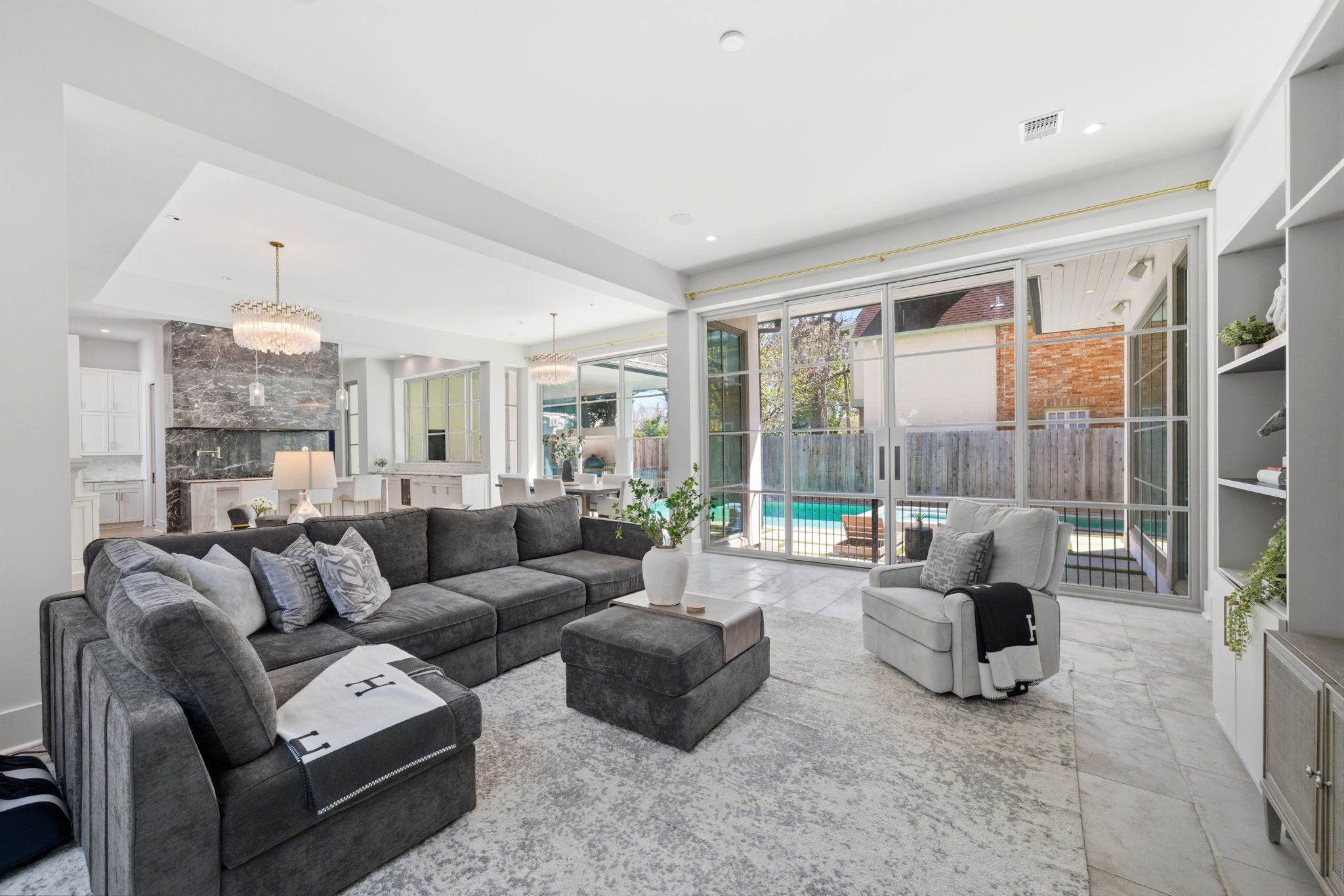 2615 Yupon Street Houston, TX 77006 - Photo 3 of 50 Expansive floor-to-ceiling steel and glass doors create a seamless transition to the backyard oasis, highlighting the sophisticated indoor-outdoor flow of this luxury estate. The open-concept living area features a stone-clad linear fireplace, designer chandeliers, and soaring ceilings that overlook the heated pool and sleek interior dining spaces.