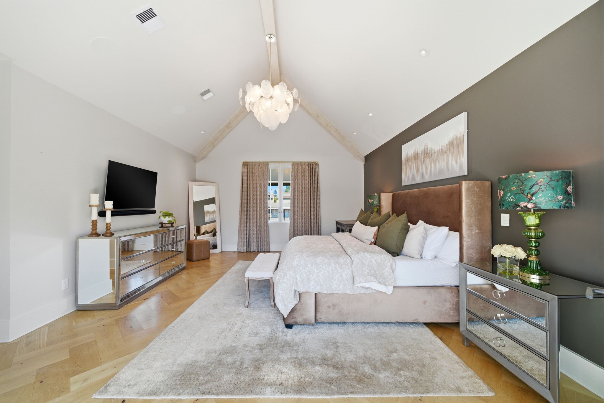 2615 Yupon Street Houston, TX 77006 - Photo 31 of 50 Vaulted ceilings with a rustic wood beam and a striking shell chandelier crown this expansive primary suite, where wide-plank herringbone white oak floors add timeless luxury. A charcoal accent wall creates a sophisticated backdrop for the king-sized bed, while a mirrored dresser and wall-mounted media center enhance the contemporary aesthetic. Floor-to-ceiling glass doors offer easy access to a private balcony overlooking the lush yard, flooding the room with natural light.