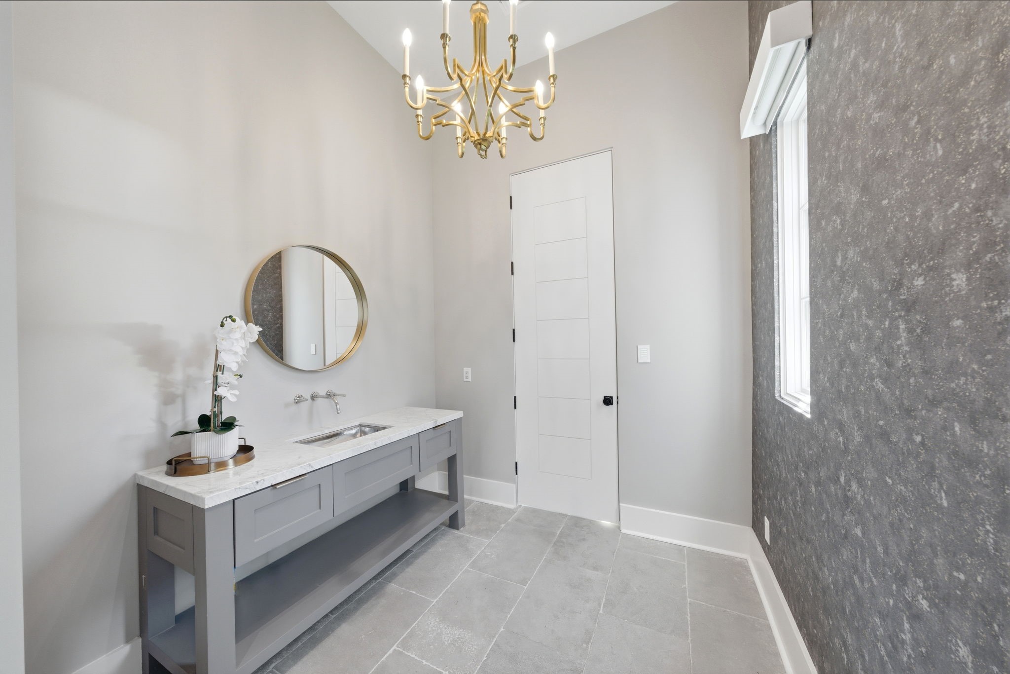 2615 Yupon Street Houston, TX 77006 - Photo 32 of 50 Elevated by high ceilings and a tiered gold chandelier, this sophisticated pool bath features a sleek gray vanity with a wide marble countertop and a modern trough-style sink. Textured charcoal wallpaper creates a bold accent wall against soft neutral tones and large-format stone tile flooring. A clean five-panel door and round gold-framed mirror complete the designer finishes, providing a refined and convenient transition from the outdoor oasis.