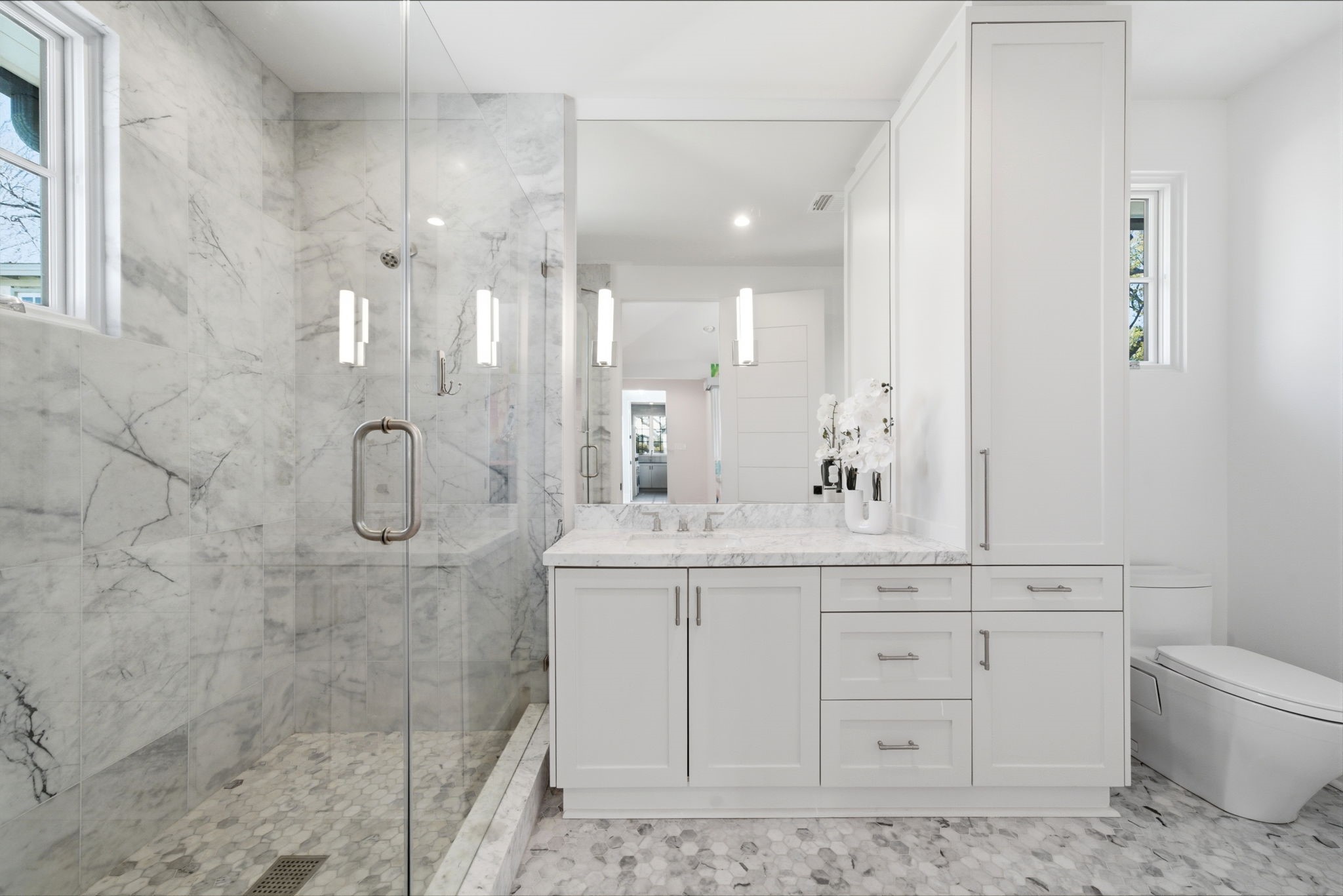 2615 Yupon Street Houston, TX 77006 - Photo 34 of 50 Spa-inspired ensuite bathroom features a glass-enclosed shower with floor-to-ceiling marble tile and a modern hexagonal mosaic floor. The crisp white custom vanity is topped with a thick marble counter and a trough-style sink, while a matching floor-to-ceiling linen cabinet provides expansive storage. High-end designer fixtures, a large frameless mirror, and a sleek low-profile commode complete this contemporary 2022 custom estate retreat.