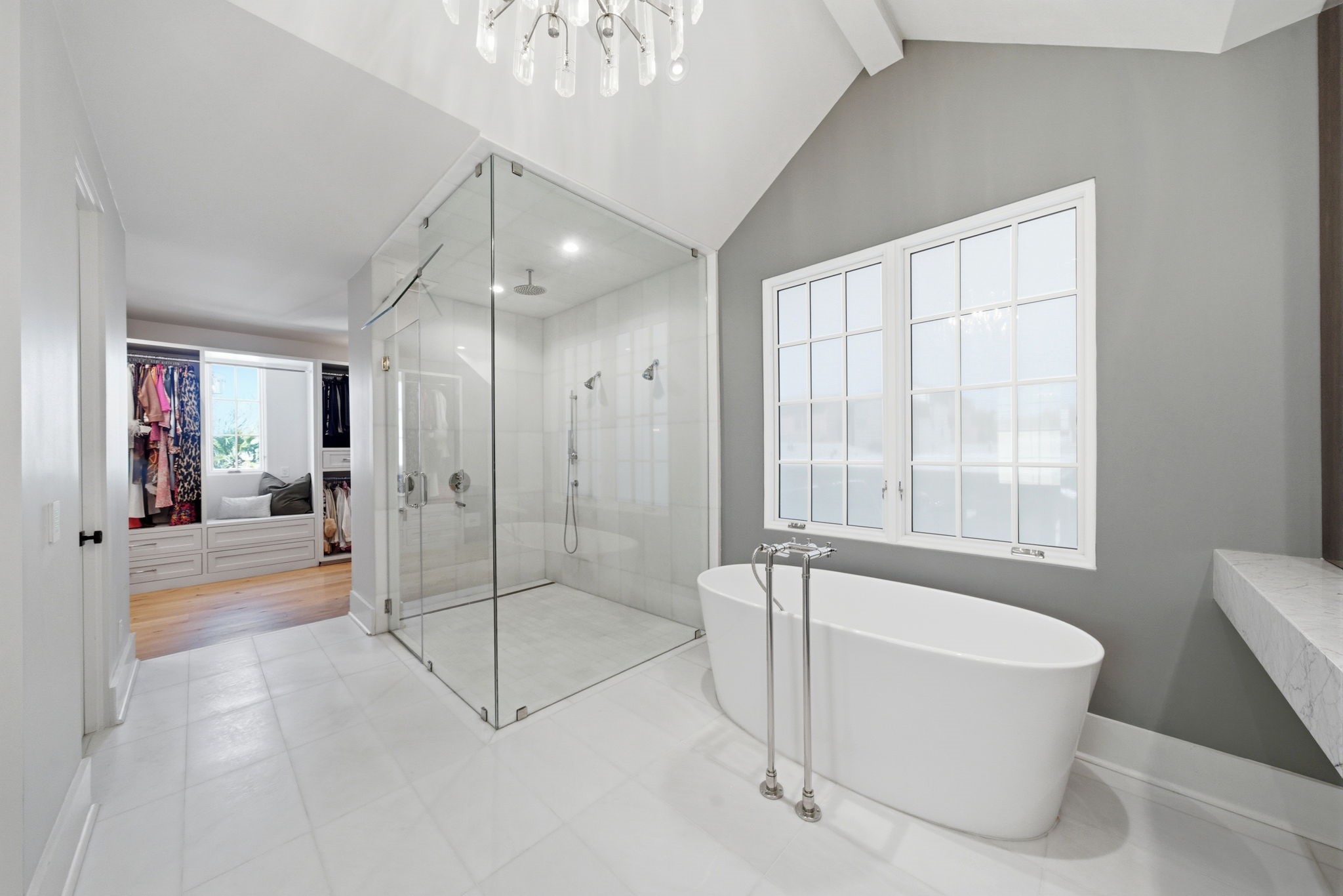 2615 Yupon Street Houston, TX 77006 - Photo 39 of 50 Spa-inspired primary ensuite bathroom features a freestanding soaking tub positioned beneath a large gridded window, offering a serene retreat within this 2022 custom estate. A massive glass-enclosed shower with multiple showerheads and floor-to-ceiling tile stands adjacent to the vaulted ceilings and designer chandelier. The open layout flows seamlessly into an expansive walk-in closet with custom built-ins and a sunlit window seat.