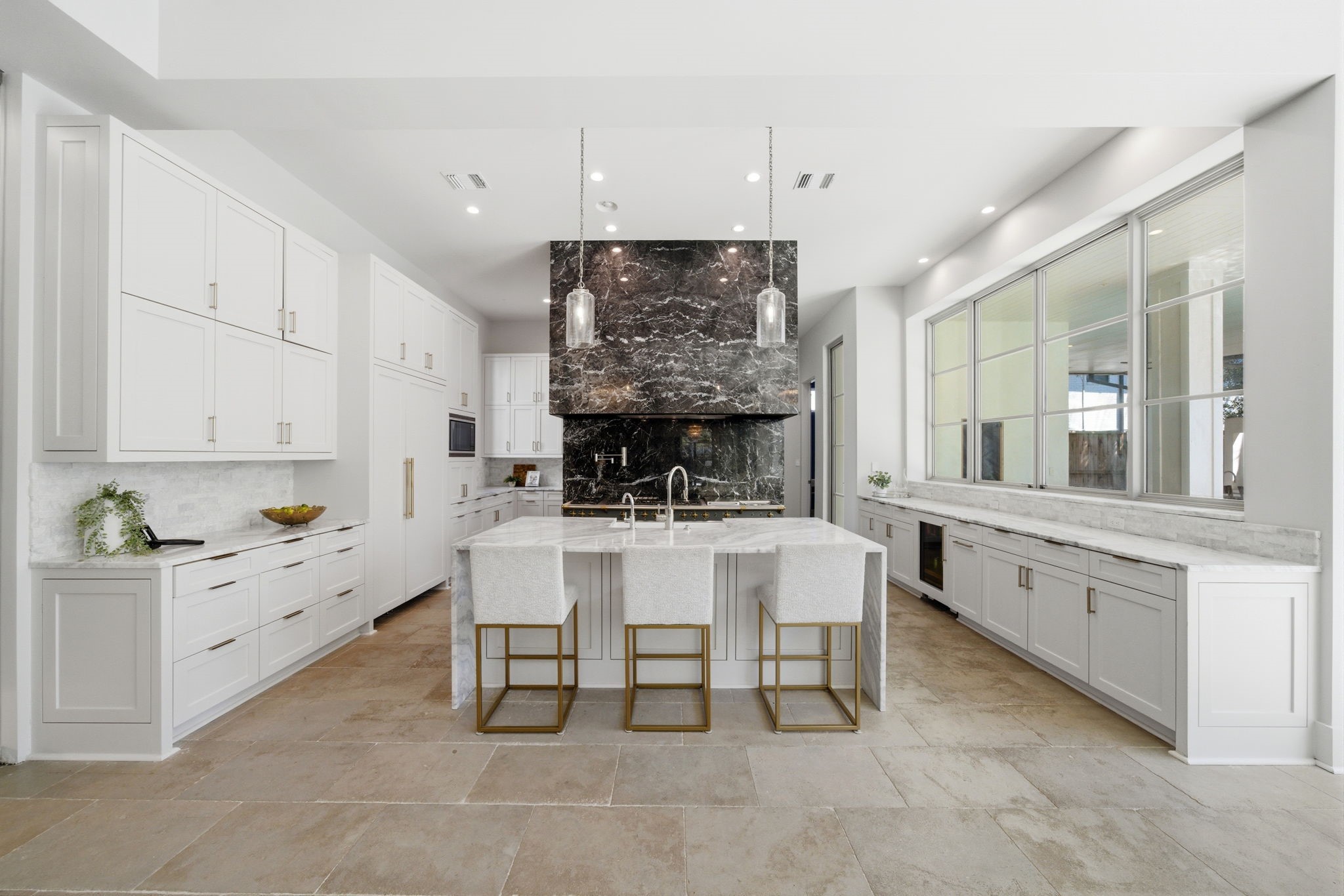 2615 Yupon Street Houston, TX 77006 - Photo 4 of 50 Gourmet excellence defines this kitchen, featuring a striking black marble range hood and backsplash that serves as a bold centerpiece against crisp white custom cabinetry. The oversized marble island provides ample seating and professional-grade functionality, while expansive windows offer serene courtyard views. Above, designer pendant lighting and high ceilings enhance the airy, open-concept layout.