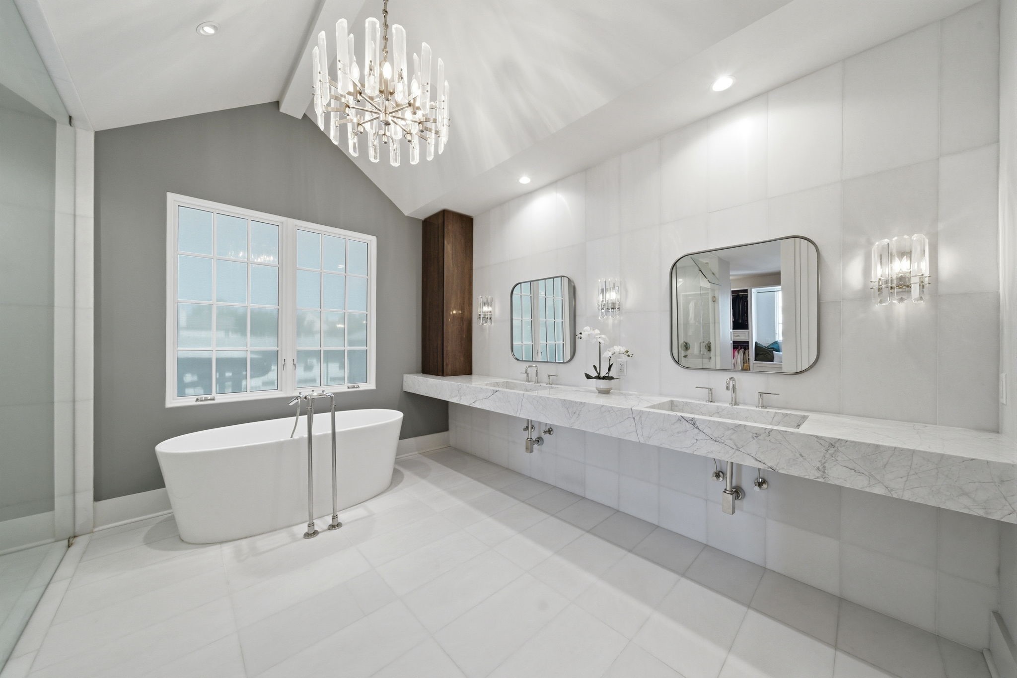 2615 Yupon Street Houston, TX 77006 - Photo 41 of 50 Vaulted ceilings and a cascading crystal chandelier crown this spa-inspired primary ensuite, anchored by a sleek freestanding soaking tub. An expansive marble-topped floating vanity spans the length of the room, featuring dual trough sinks and designer sconce lighting. Natural light from the gridded window and a soft grey accent wall complete this luxurious 2022 custom estate retreat.
