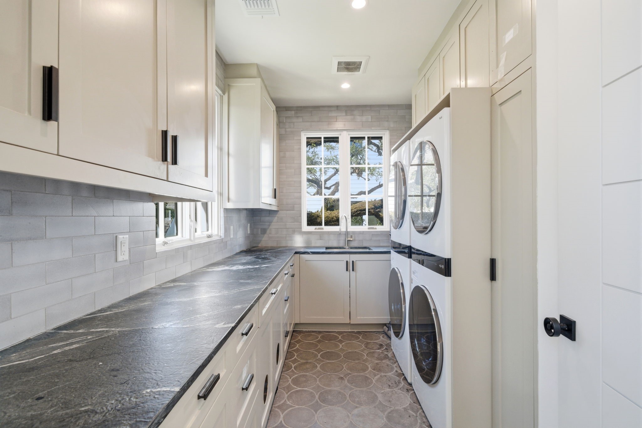 2615 Yupon Street Houston, TX 77006 - Photo 42 of 50 Large gridded window frames a majestic oak tree, filling this high-end laundry room with natural light and a sense of serenity. Premium white cabinetry with sleek black hardware offers expansive storage, while the dark stone countertops provide a sophisticated folding area. Dual stacked washers and dryers ensure maximum efficiency, complemented by a deep utility sink, designer tile flooring, and a subway tile backsplash that reaches the ceiling.