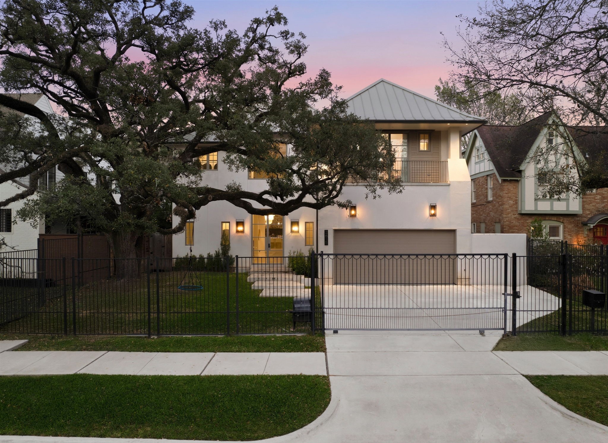 2615 Yupon Street Houston, TX 77006 - Photo 44 of 50 Stately oak trees and professional landscaping frame 2615 Yupon St, highlighting a striking stucco facade and sleek metal roof. A secure black iron gate and private driveway lead to the 3-car garage, while designer lantern-style sconces illuminate the floating concrete walkway. This 2022 contemporary estate commands exceptional curb appeal with clean architectural lines and a private balcony.