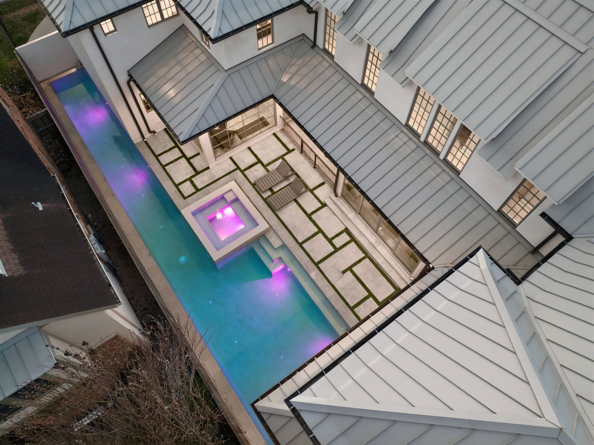 2615 Yupon Street Houston, TX 77006 - Photo 45 of 50 Aerial perspective showcases the striking L-shaped architecture of this 2022 custom estate, centered around a linear heated pool and an elevated square spa. Vibrant LED accent lighting illuminates the turquoise water, while sleek geometric pavers with turf inlays create a sophisticated path between the home gym and the outdoor lounging deck. Metal roofing and expansive gridded windows emphasize the contemporary luxury of this private courtyard oasis.