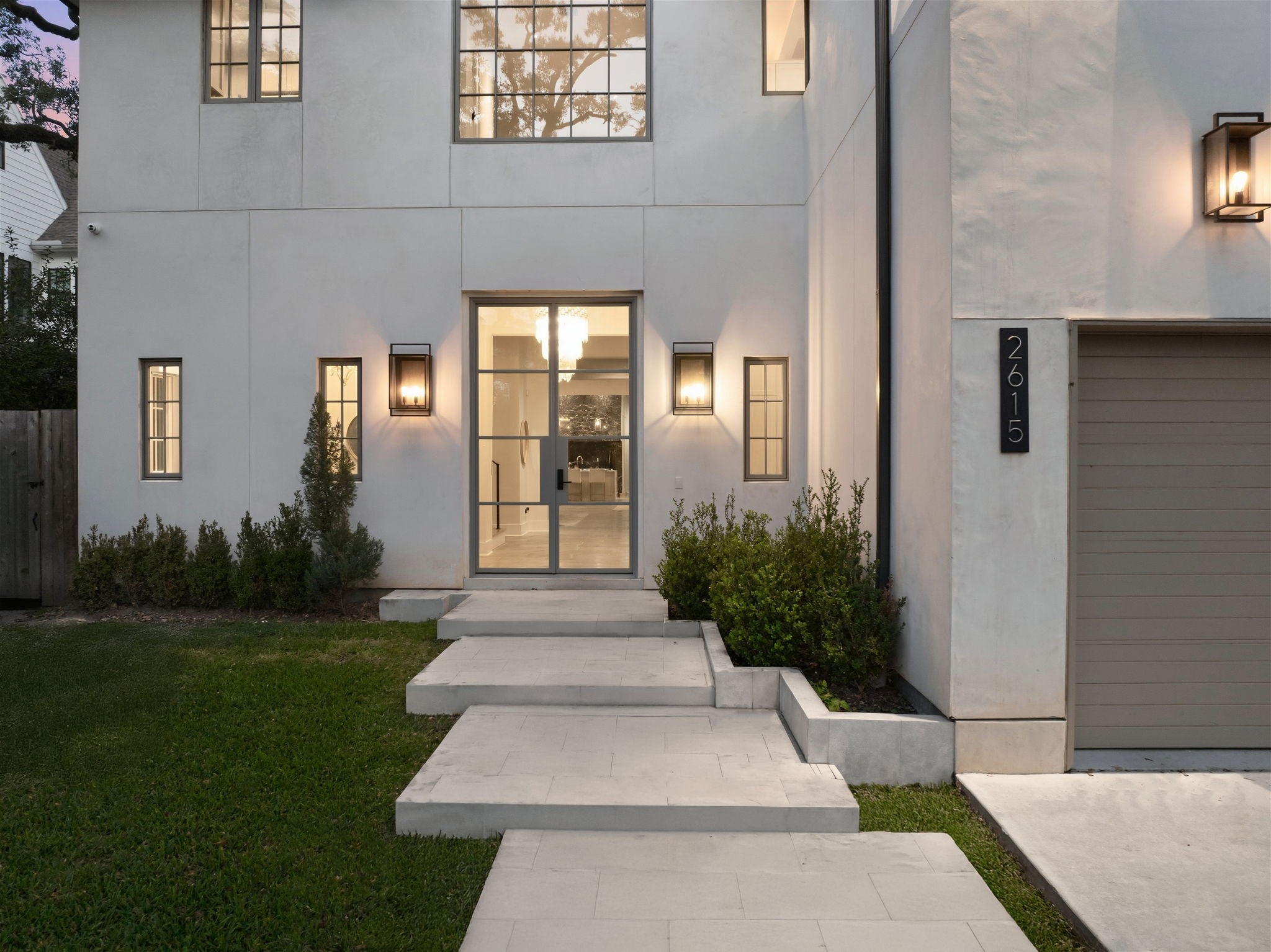 2615 Yupon Street Houston, TX 77006 - Photo 8 of 50 Modern lantern-style sconces flank the steel and glass entry door, creating a sophisticated glow against the smooth stucco exterior. Floating concrete steps lead to the grand foyer, where a designer chandelier is visible through the expansive windows. This 2022 custom estate at 2615 Yupon St blends sleek architectural lines with lush landscaping for an inviting first impression.
