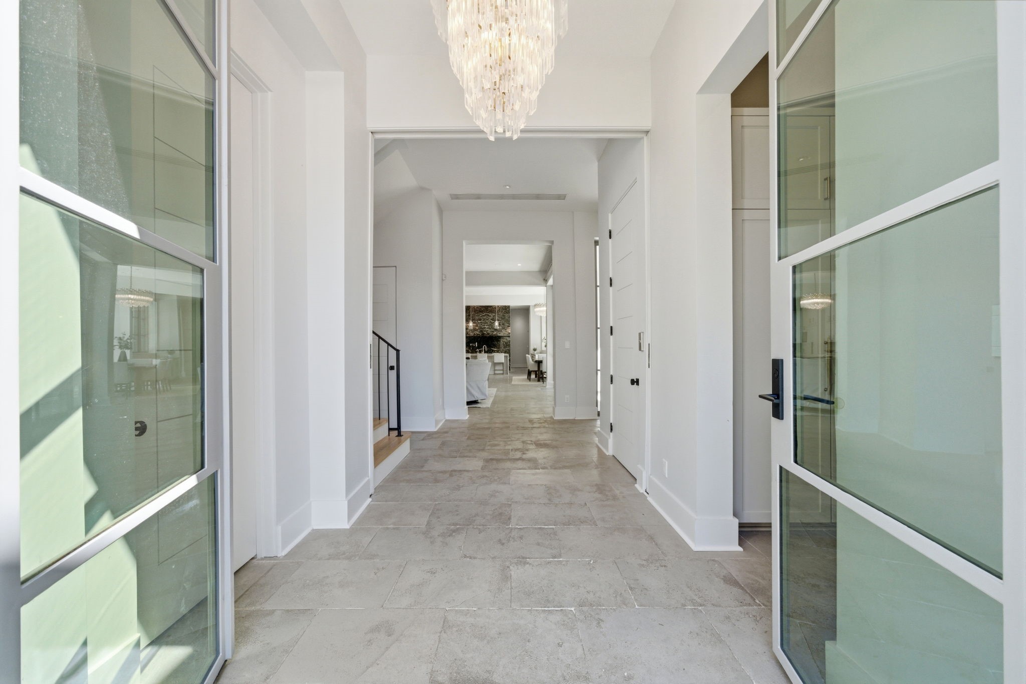 2615 Yupon Street Houston, TX 77006 - Photo 9 of 50 Grand foyer features elegant architectural details, including a cascading crystal chandelier and durable stone tile flooring. Steel and glass doors offer glimpses into the dedicated study and mudroom, while the wide hallway flows effortlessly toward the light-filled main entertainment areas. Refined neutral tones and clean lines define this 2022 custom estate’s sophisticated entry.