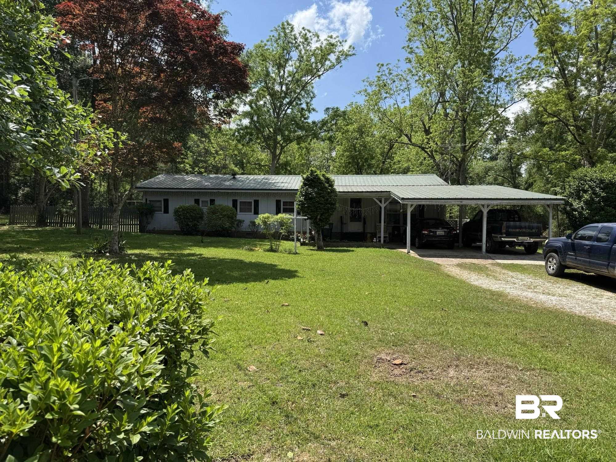 Single story home with metal roof, dirt driveway, a carport, fence, and a front lawn