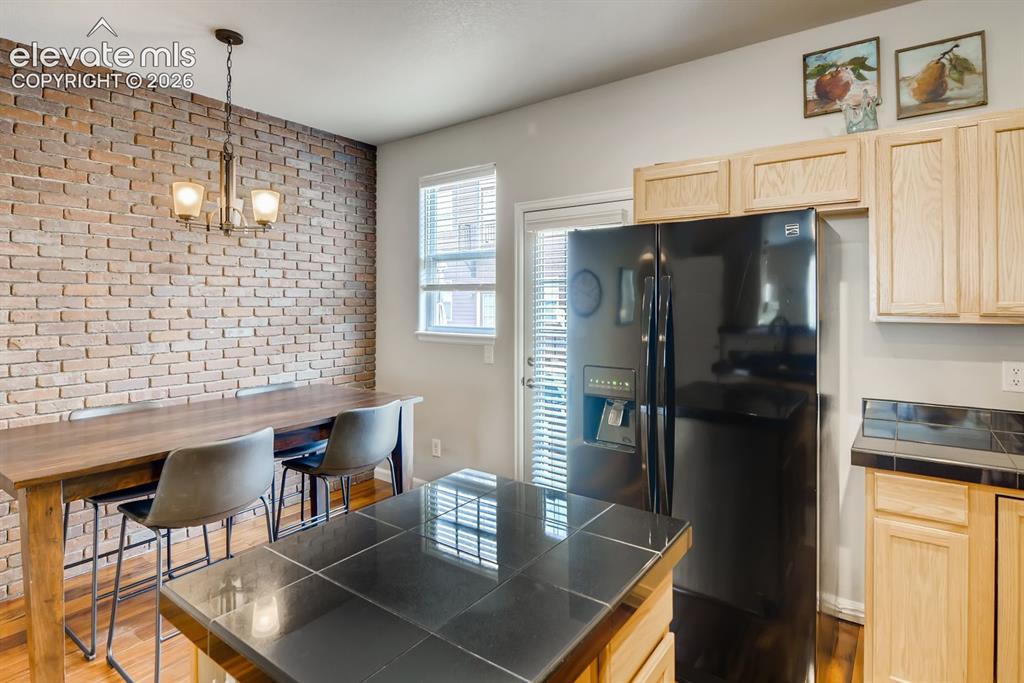 846 Victoria Ridge Point Colorado Springs, CO 80903 - Photo 11 of 27 Kitchen featuring a prominent brick accent wall, light wood cabinetry, black tile countertops, and a black side-by-side refrigerator