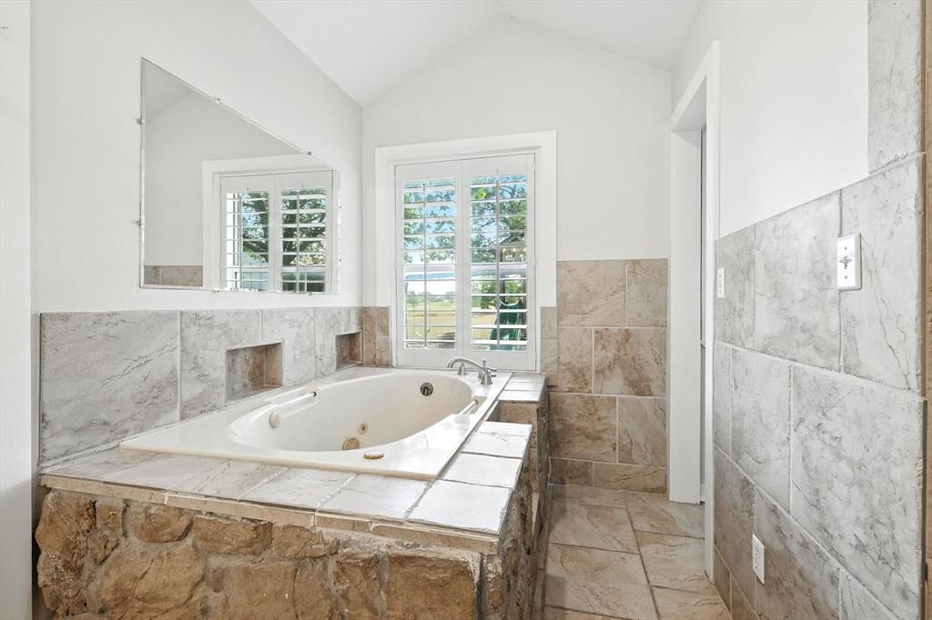 185 Quail Run Court Decatur, TX 76234 - Photo 23 of 37 Built-in jetted tub surrounded by stone and tile, featuring a mounted mirror, vaulted ceiling, and plantation-shuttered windows