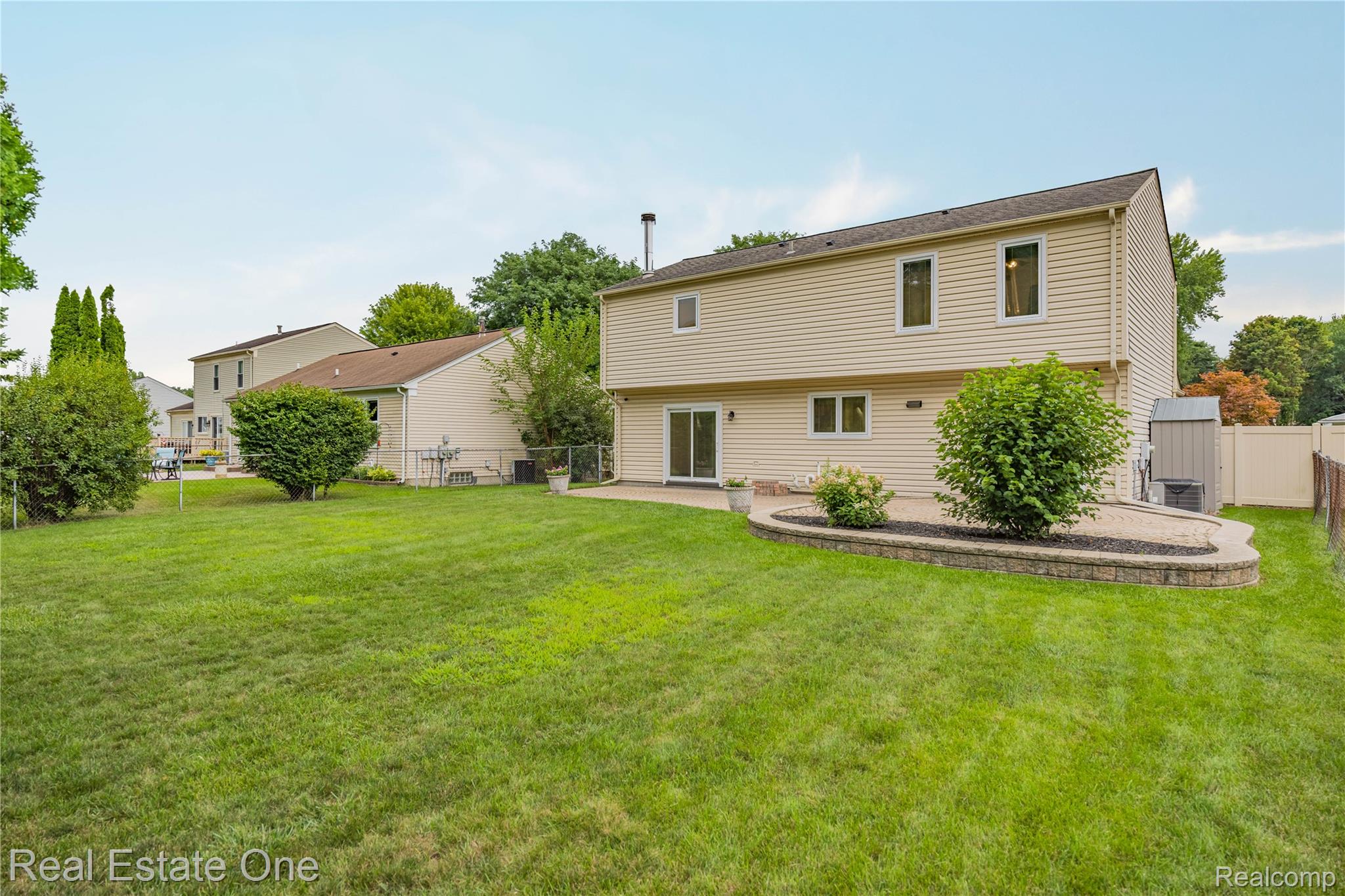 39750 Cheviot Road Canton, MI 48188 - Photo 60 of 71 Expansive green lawn with mature landscaping, a raised garden bed with stone retaining wall, and a private patio with sliding glass door access
