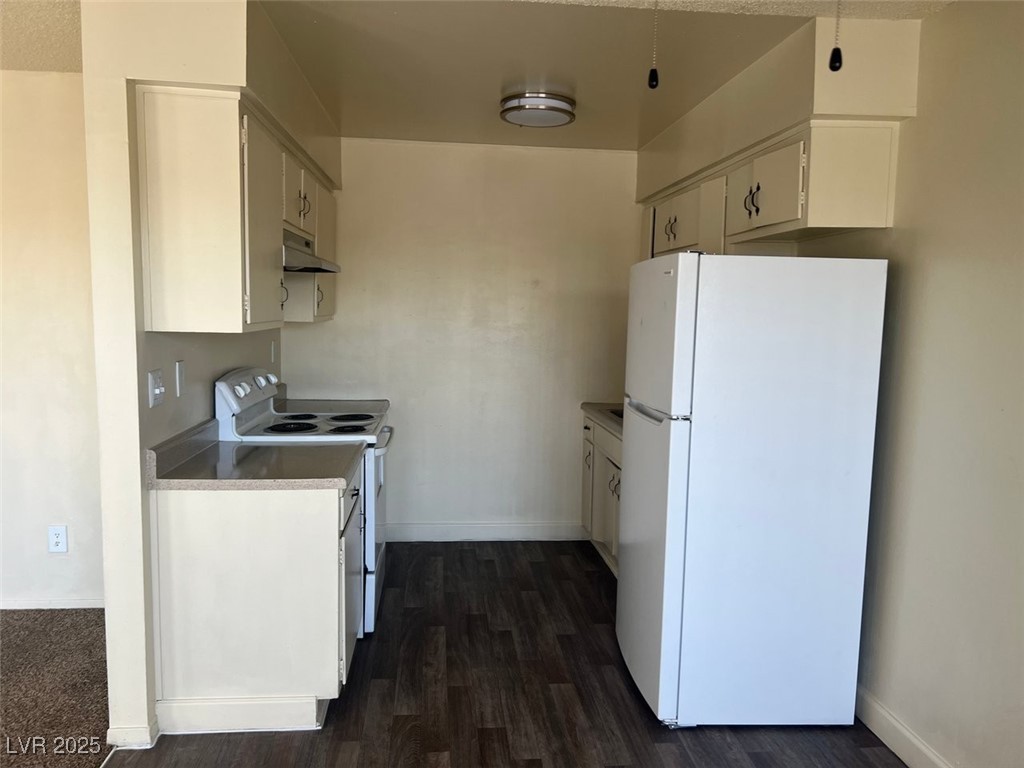 Kitchen with white appliances, under cabinet range hood, dark wood finished floors, and baseboards