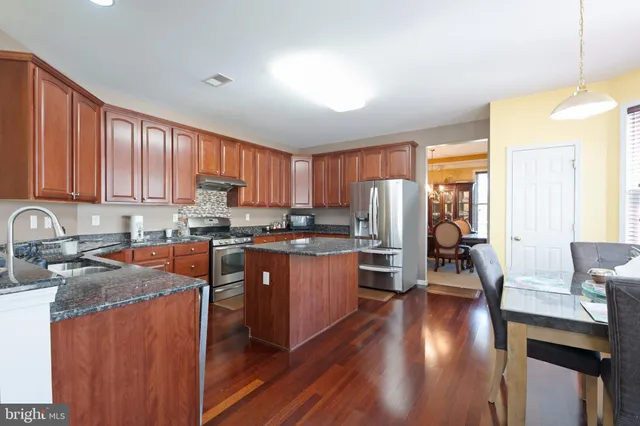 a kitchen with stainless steel appliances granite countertop wooden cabinets a refrigerator a sink a stove a dining table and chairs with wooden floor
