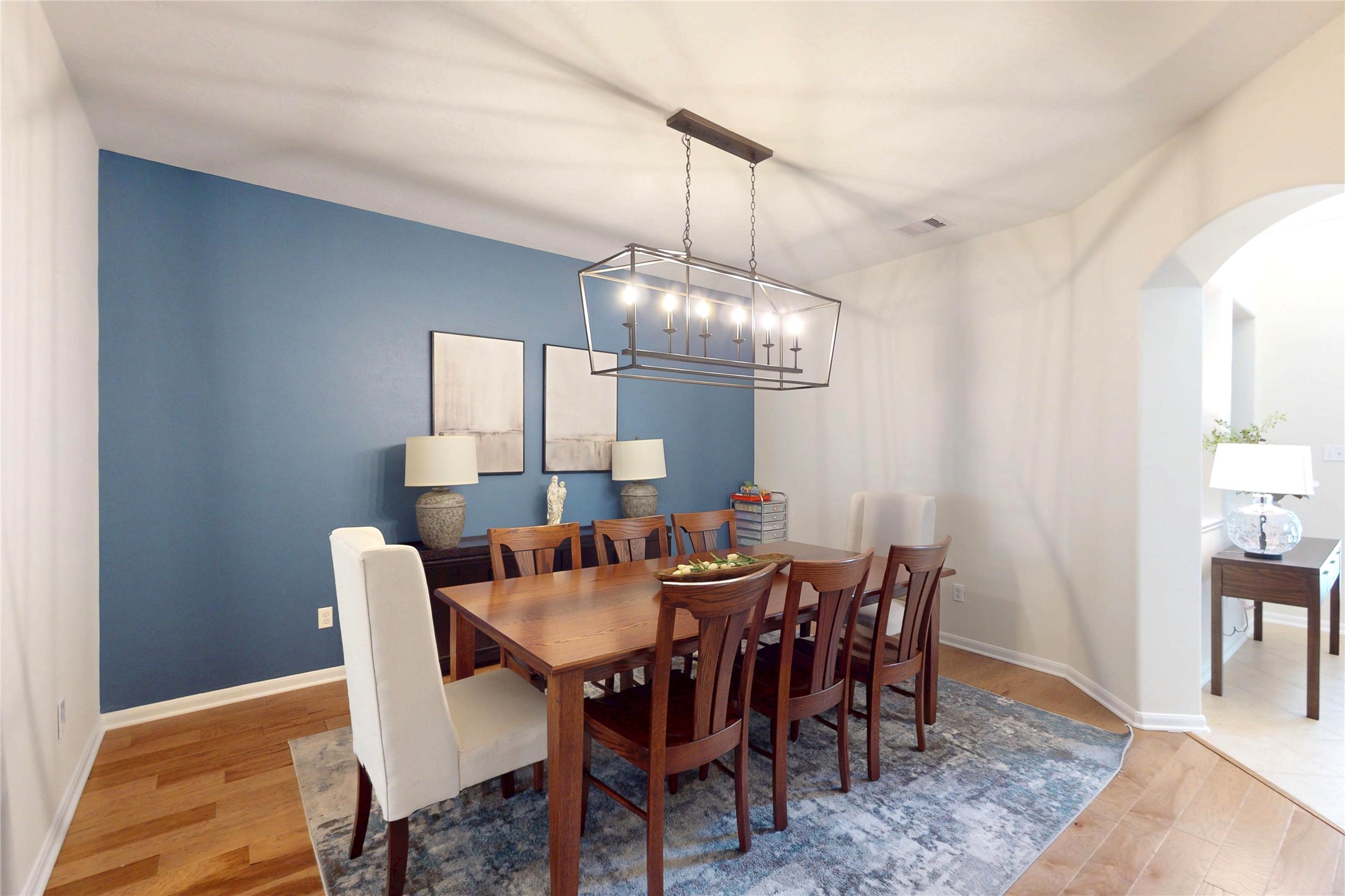 17402 Rainer Valley Lane Humble, TX 77346 - Photo 4 of 33 This dining room features a stylish blue accent wall, a large wooden table with seating for eight, and a modern chandelier. It has hardwood flooring and an adjacent archway leading to another room, creating an open and inviting space.