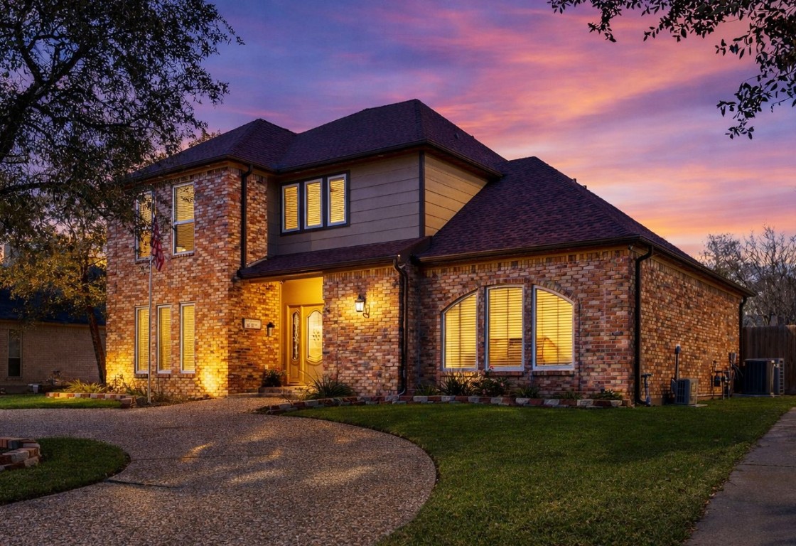 This charming two-story brick home features a beautifully lit exterior with large arched windows and a welcoming front entrance. The property includes a curved driveway and is set against a picturesque sunset, offering an inviting and elegant curb appeal.